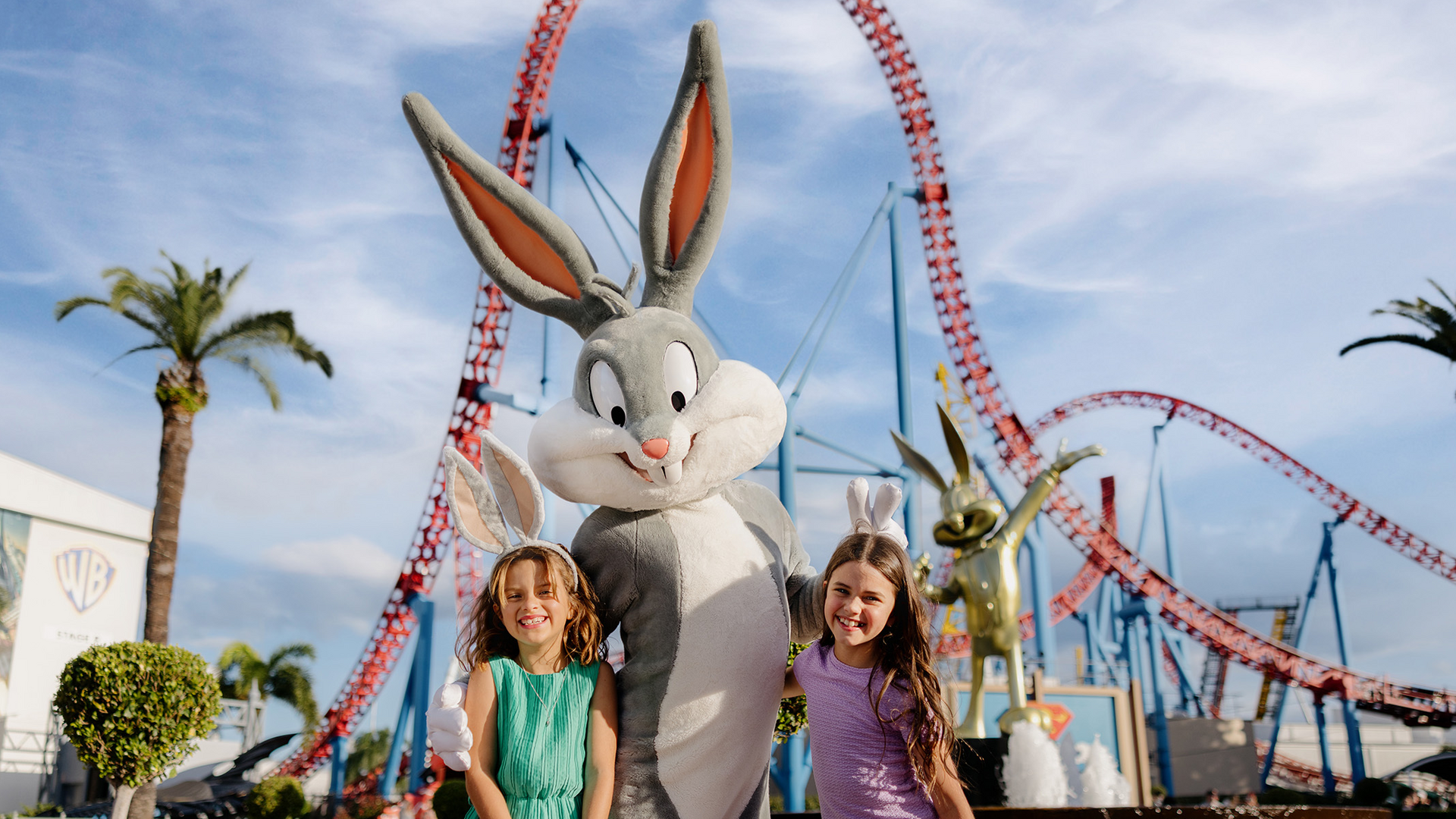 Two smiling children stand next to a person dressed as Bugs Bunny at an amusement park, with a large roller coaster and palm trees in the background under a blue sky.
