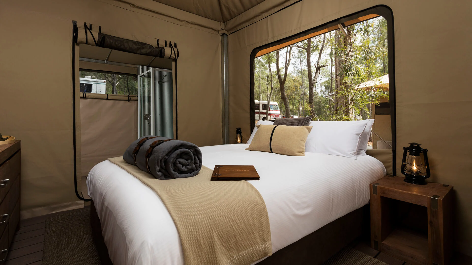 A neatly made bed with beige linens and rolled towels sits in a canvas tent room with a window showing trees outside; a small bedside table holds a lantern.