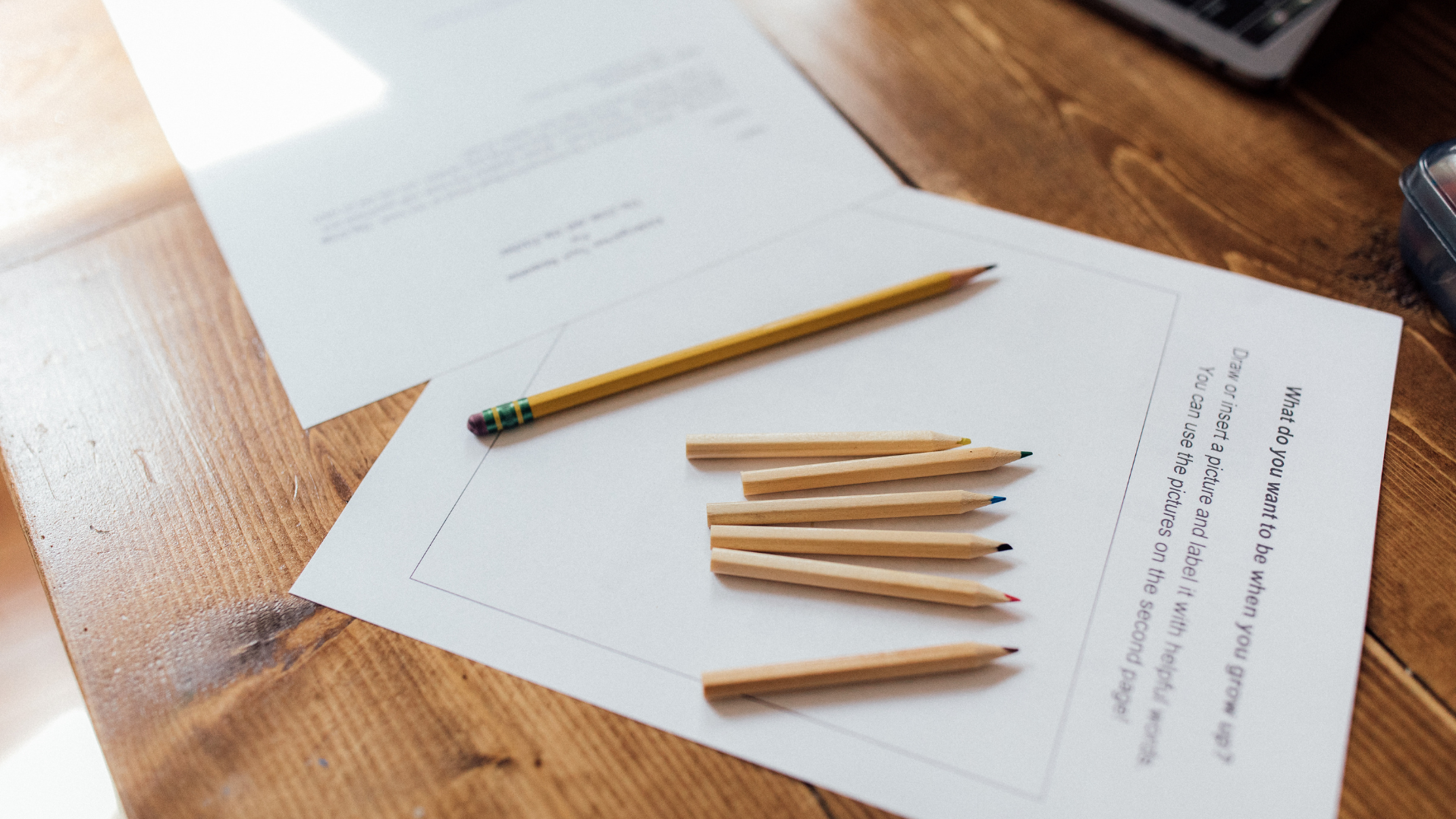 A close-up of several colored pencils and papers on a wooden table, with soft sunlight shining across the scene.