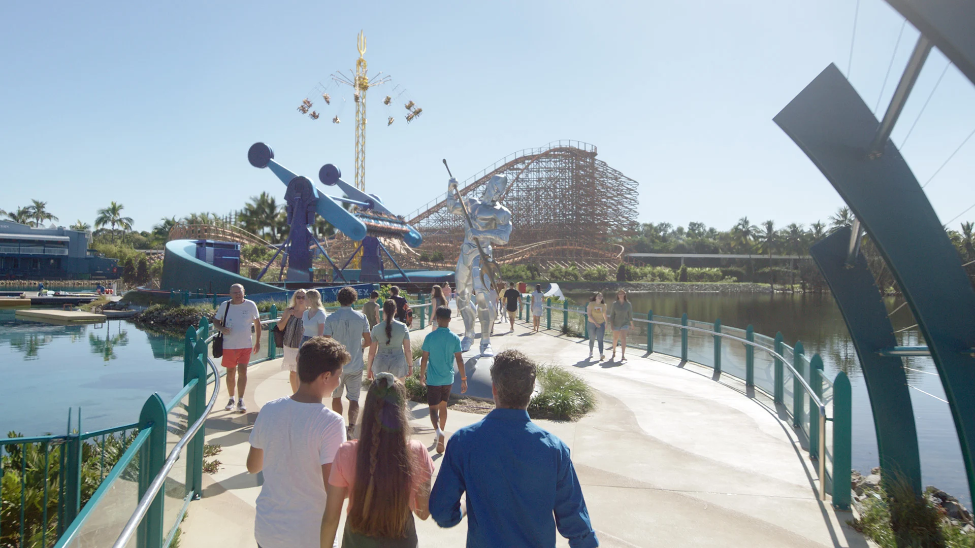 People walk along a path toward a roller coaster and amusement park rides on a sunny day, passing a silver statue near a pond.