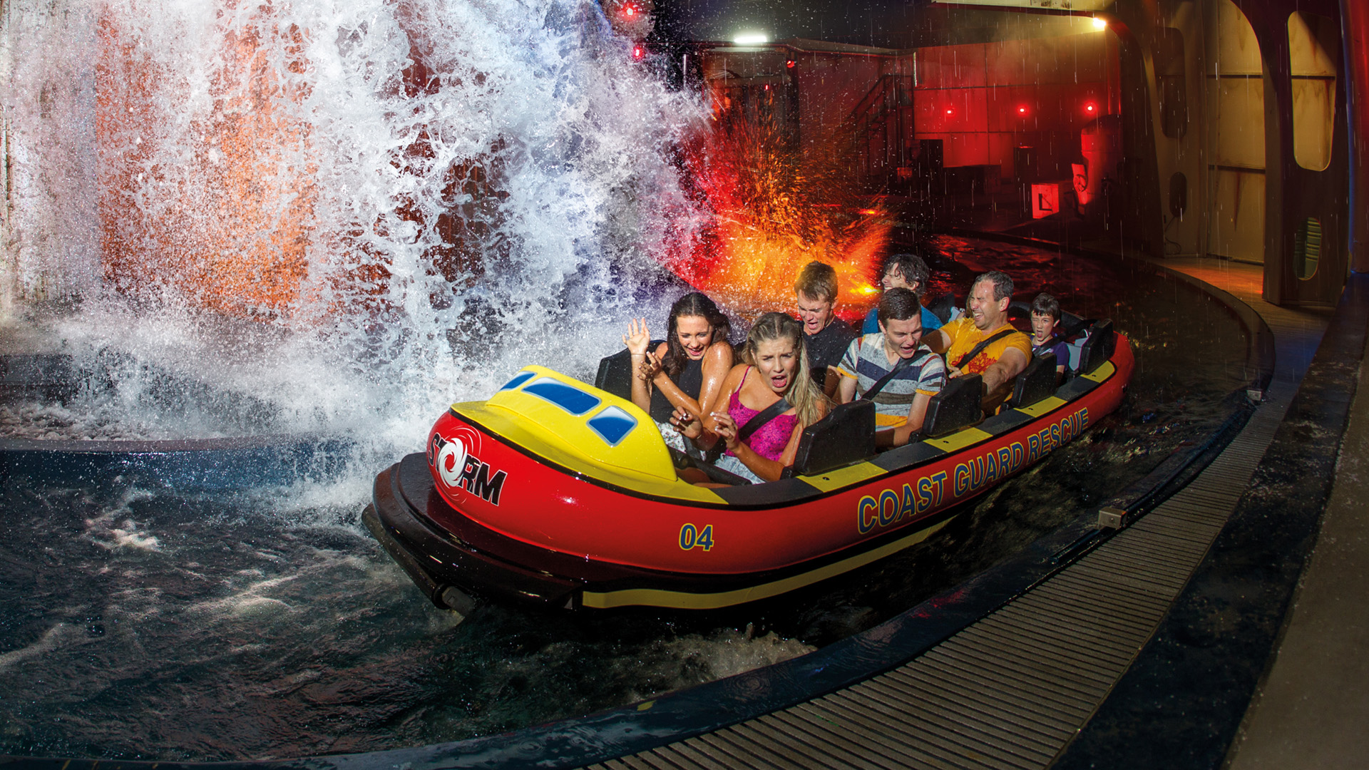 A group of people, including children and adults, ride a circular raft on a water ride indoors, passing by a large, splashing waterfall with colorful lights and water spraying all around them.