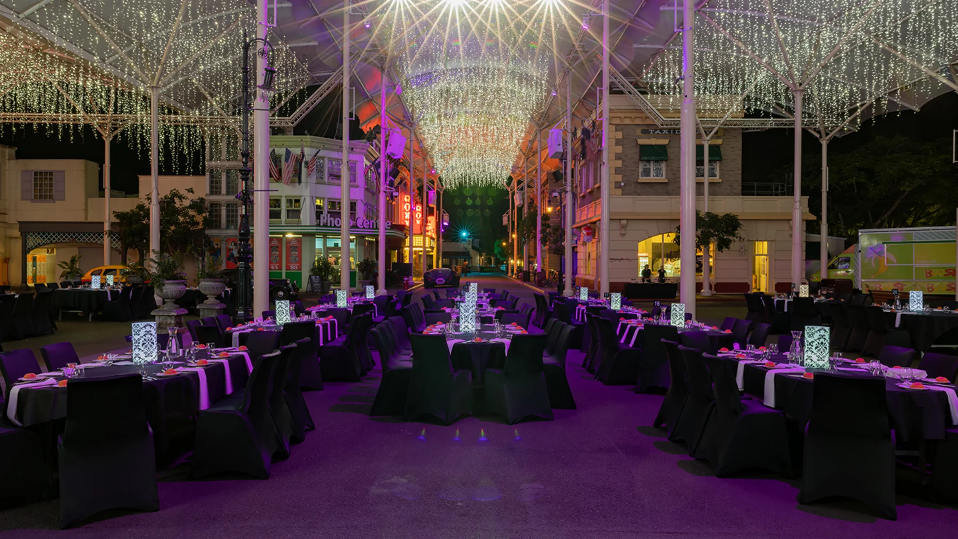 Elegant outdoor banquet setup at night, with round tables draped in black tablecloths, illuminated centerpieces, and string lights hanging above, creating a festive and sophisticated atmosphere.