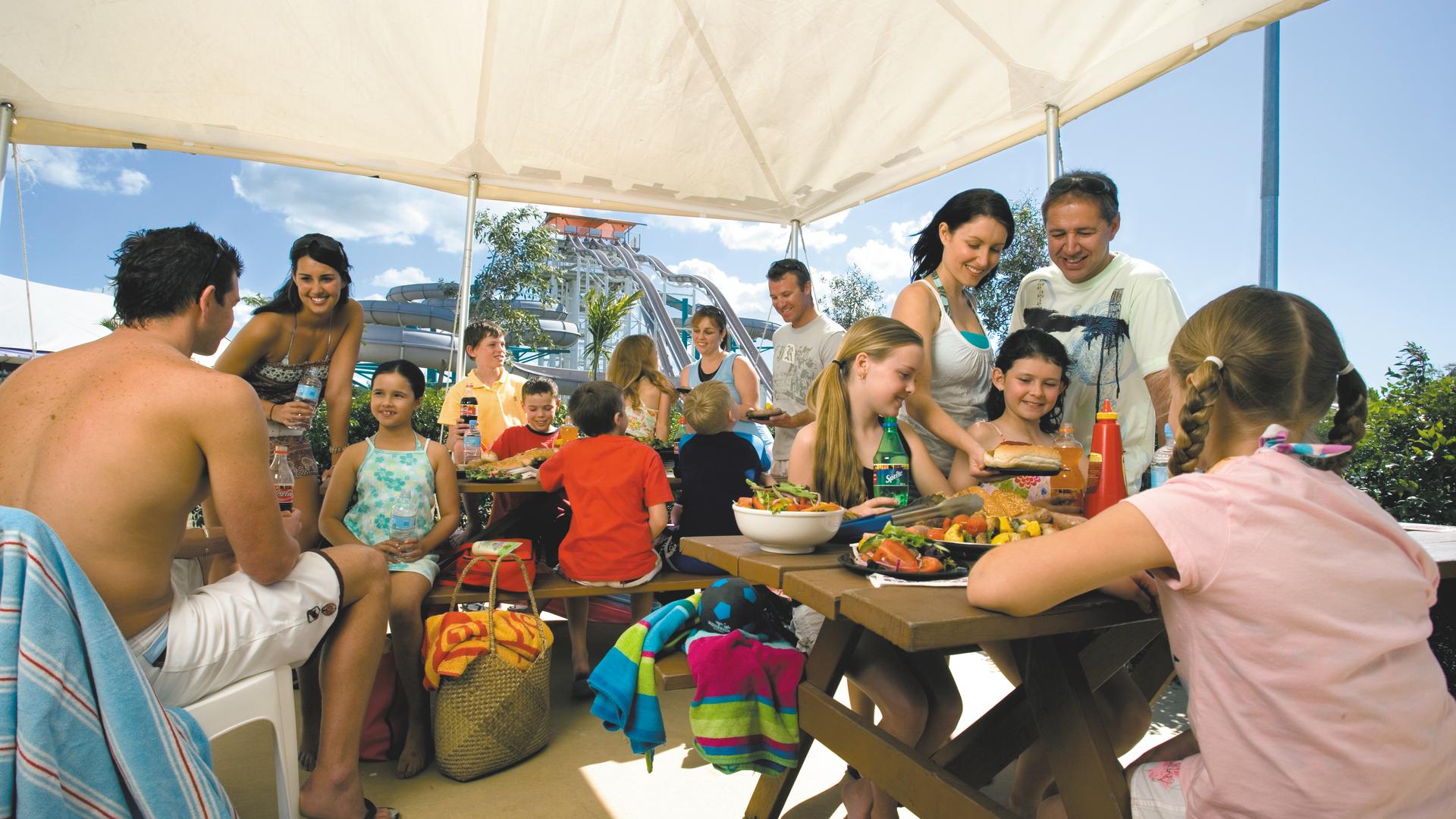 A group of adults and children gather under a canopy, enjoying food and drinks at an outdoor picnic. The atmosphere is cheerful, with people chatting and smiling. A waterslide can be seen in the background.