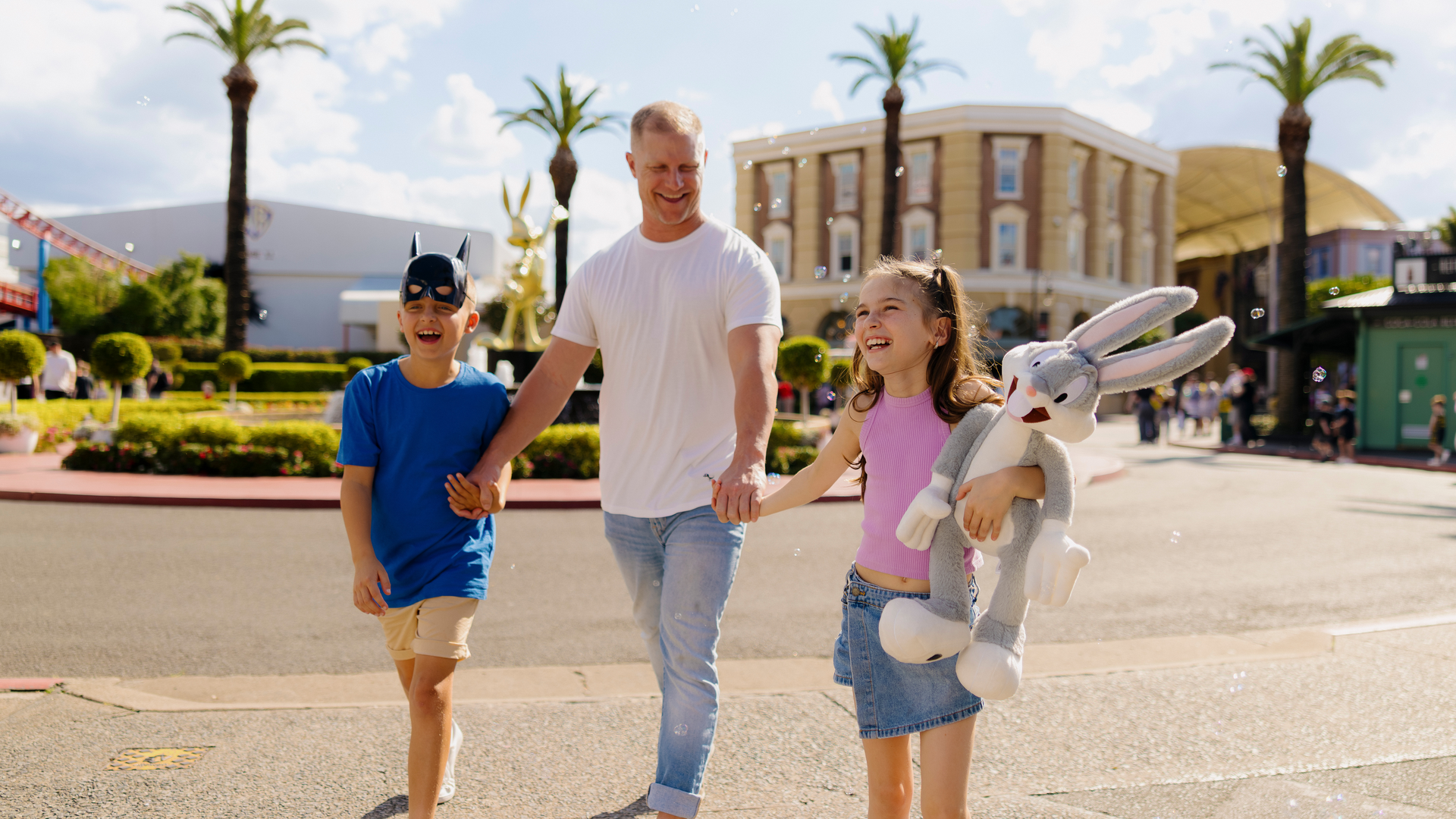 A man holds hands with a smiling boy in a Batman mask and a girl holding a large Bugs Bunny plush toy as they walk outdoors on a sunny day, with palm trees and buildings in the background.
