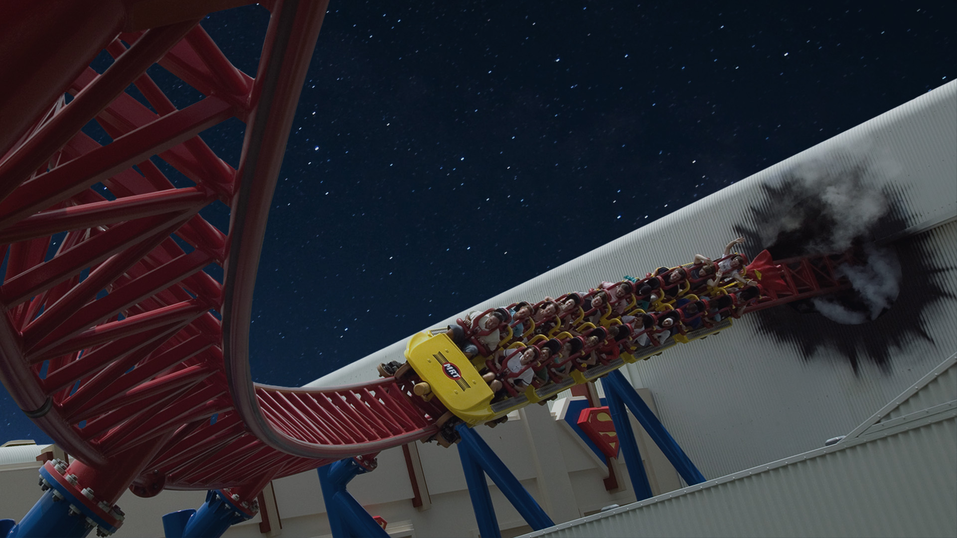 A roller coaster full of people speeds along a red track at night, with stars visible in the sky and a burst of smoke at one bend. The setting includes structures and Superman’s logo in the background.