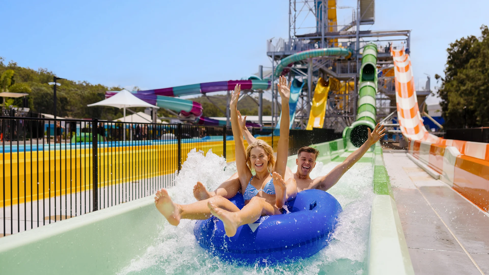 Two people ride together on a blue inflatable tube down a waterslide, splashing into the pool at the end and smiling with their arms raised. Colorful slides and structures are visible in the background.