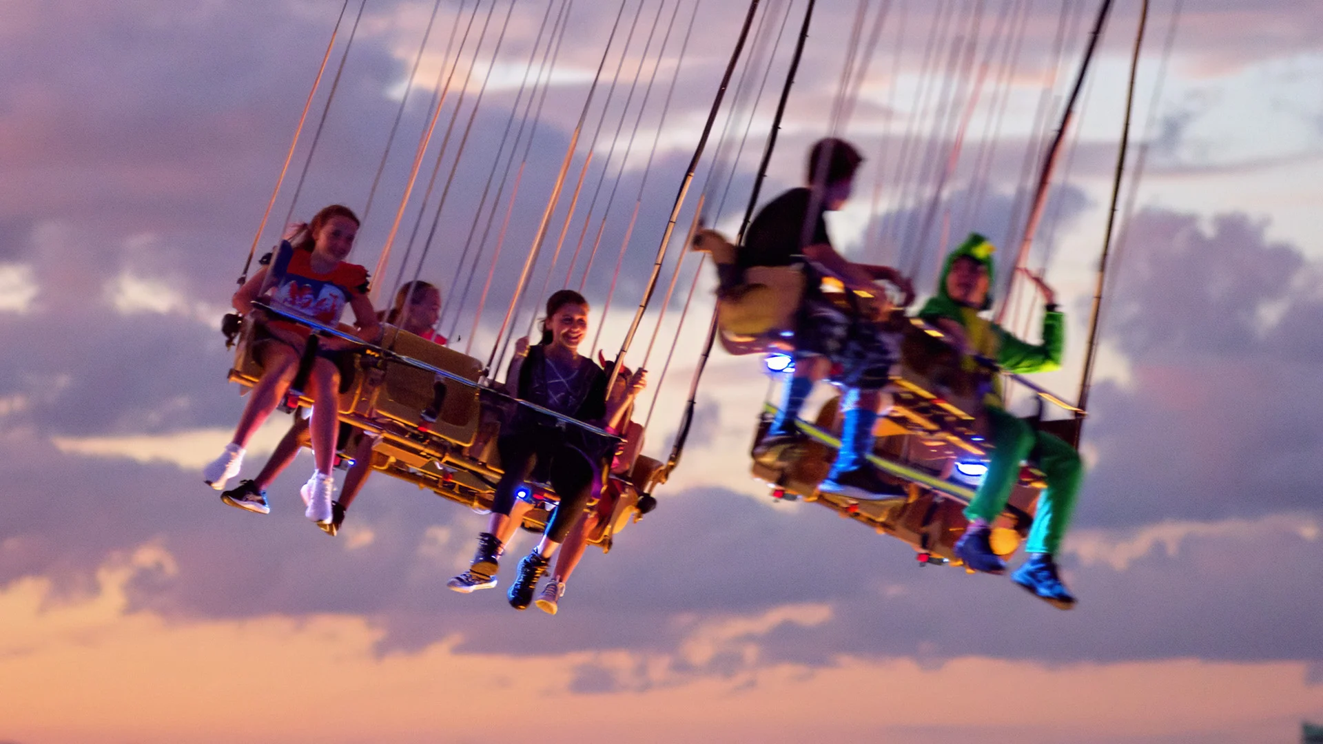 Children ride a spinning swing carousel at sunset, with clouds in the background. Some wear colorful outfits, and the ride is illuminated with soft, glowing lights.