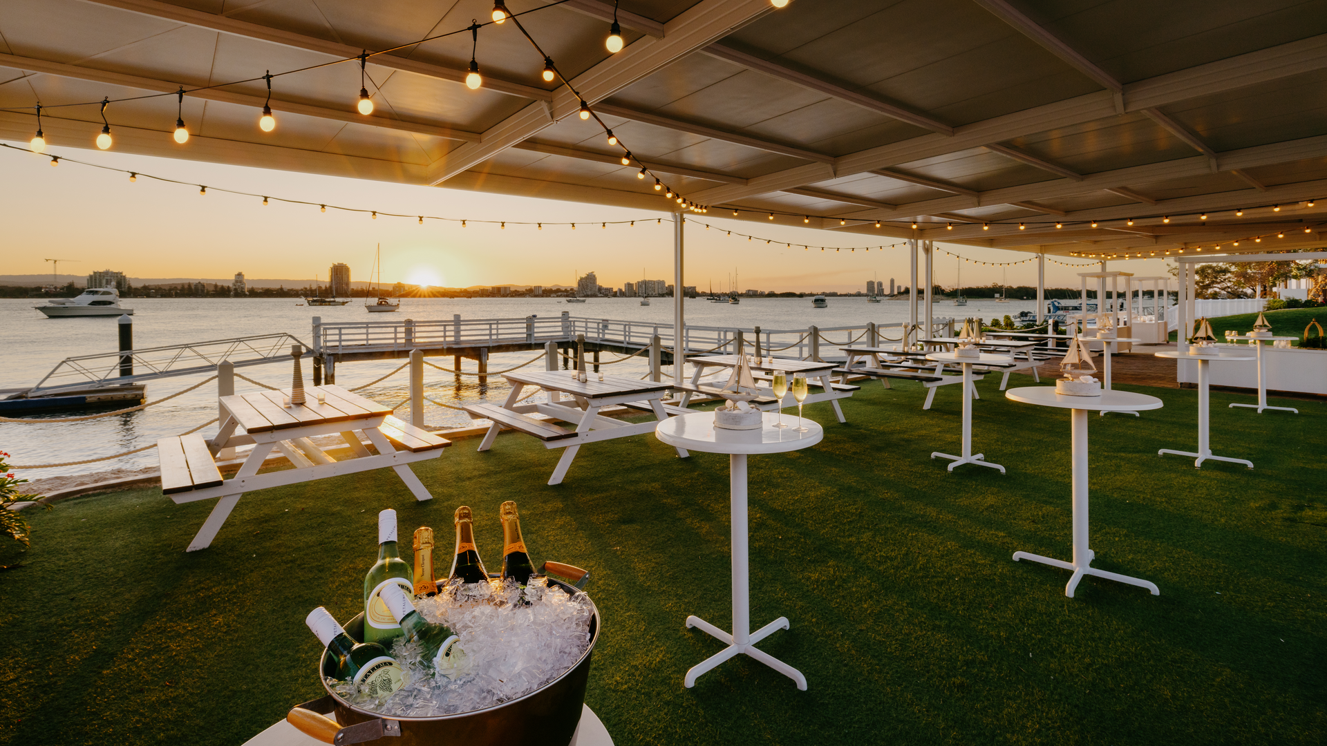 Outdoor waterfront venue at sunset with string lights, picnic tables, standing tables, and a bucket of chilled champagne bottles, overlooking a calm river with boats and city buildings in the background.