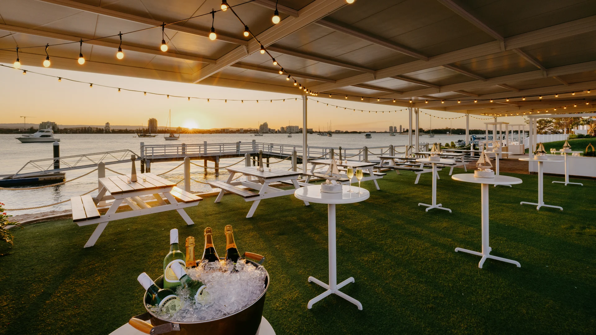 Outdoor waterfront venue at sunset with string lights, picnic tables, standing tables, and a bucket of chilled champagne bottles, overlooking a calm river with boats and city buildings in the background.