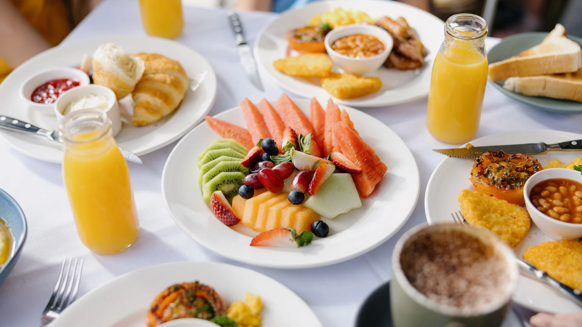 A breakfast table with plates of sliced fruit, croissants, toast, hash browns, baked beans, orange juice, and a cup of coffee.