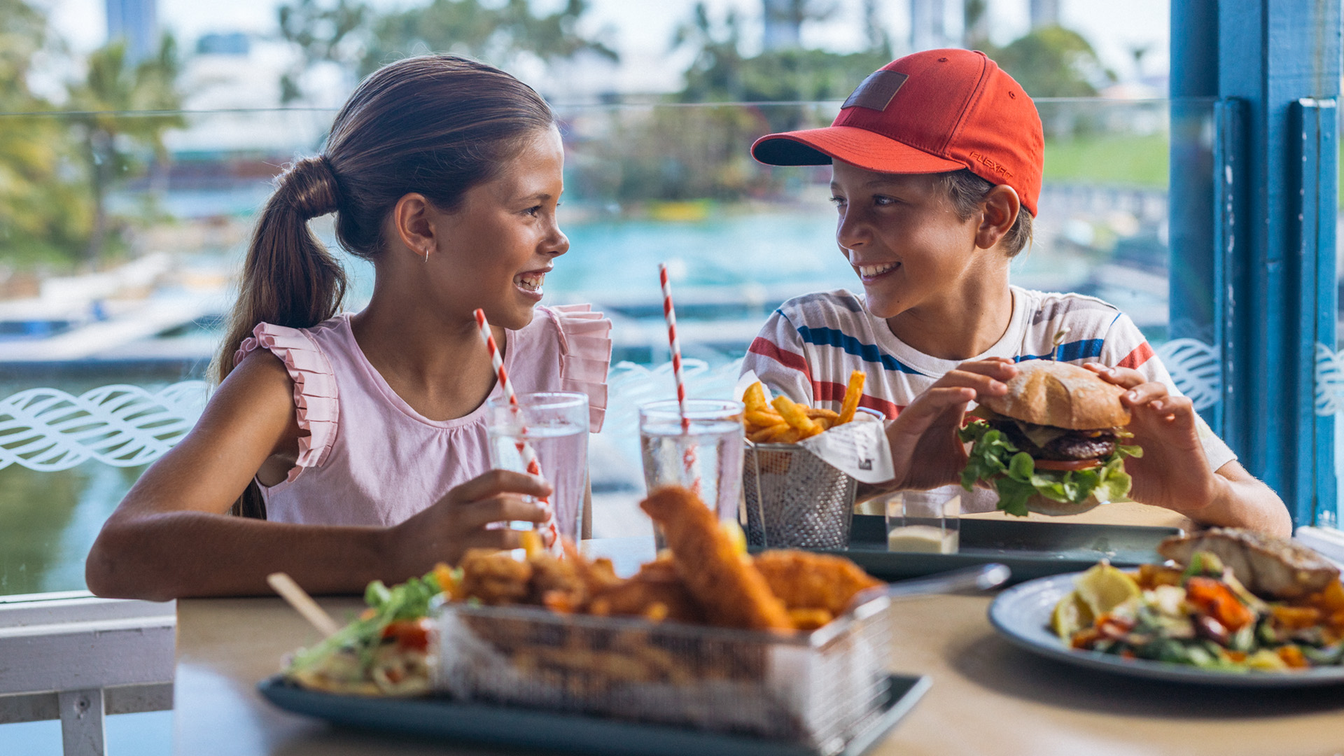 Two children sit at a table outdoors, smiling at each other while eating a meal with burgers, fries, and drinks.