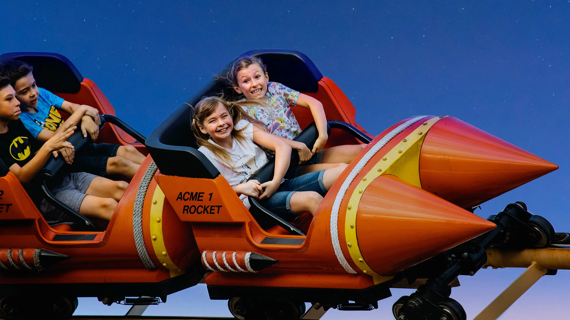 Three children smiling and holding on while riding a red and orange rocket-shaped amusement park ride against a clear blue sky.