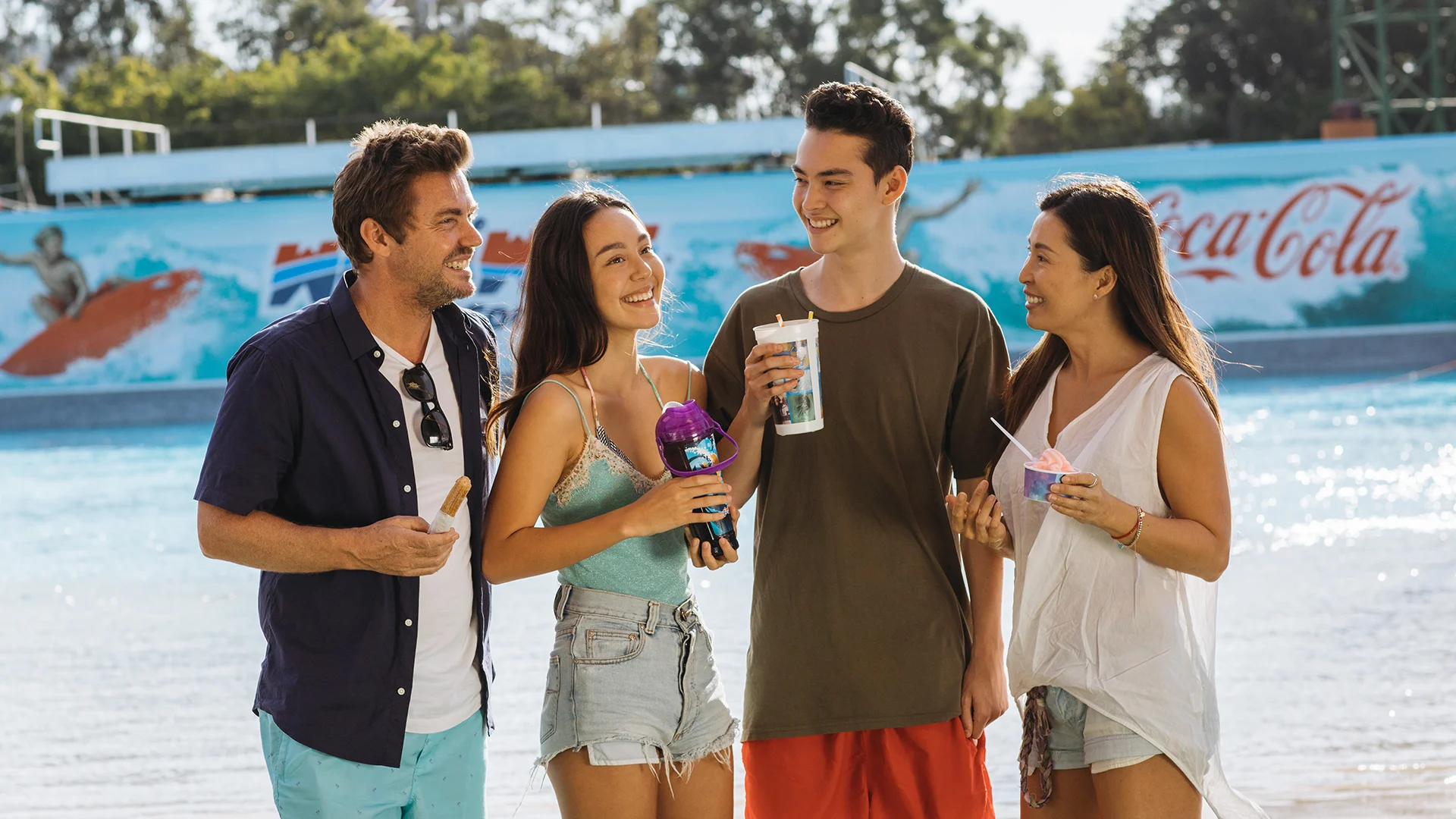 Four young adults stand together by a pool, smiling and holding drinks and snacks. A Coca-Cola advertisement and a wave pool are visible in the background. The setting appears sunny and relaxed.