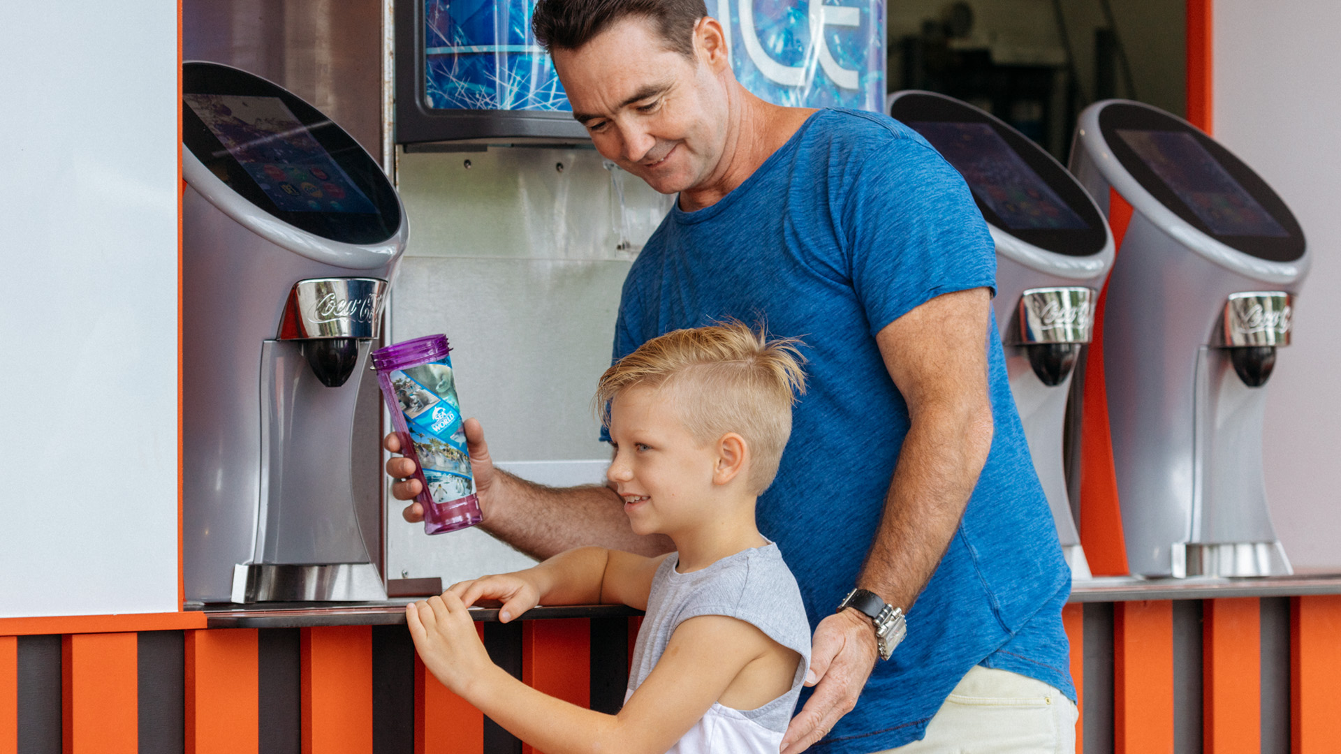 An adult helps a child fill a reusable cup at a self-serve drink machine with two other similar machines in the background.