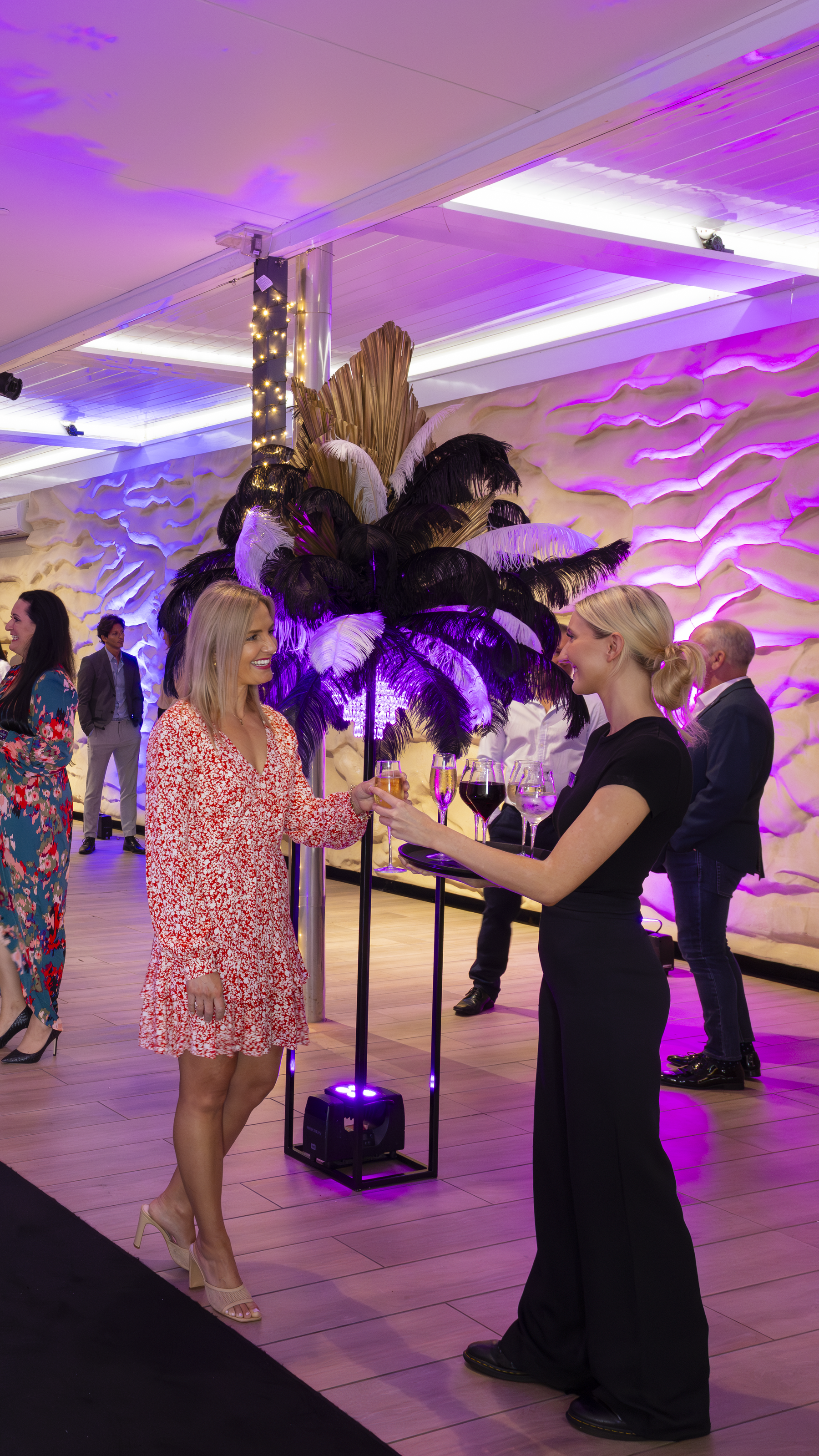 Two women chatting at an event with purple lighting and a decorative feather arrangement in the background. Other guests are mingling.