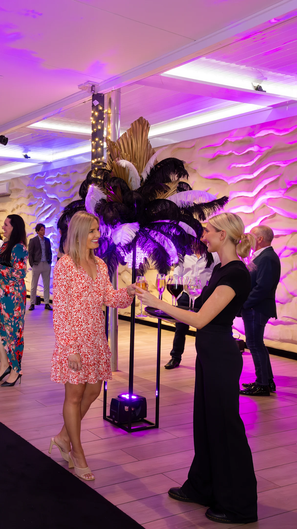 Two women chatting at an event with purple lighting and a decorative feather arrangement in the background. Other guests are mingling.