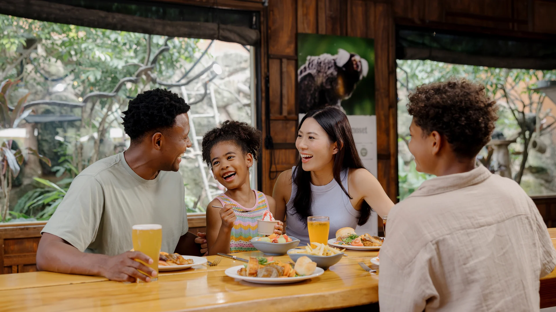 A family of four sits at a wooden table in a restaurant, smiling and enjoying food and drinks together. Large windows show lush greenery outside, creating a bright and cheerful atmosphere.