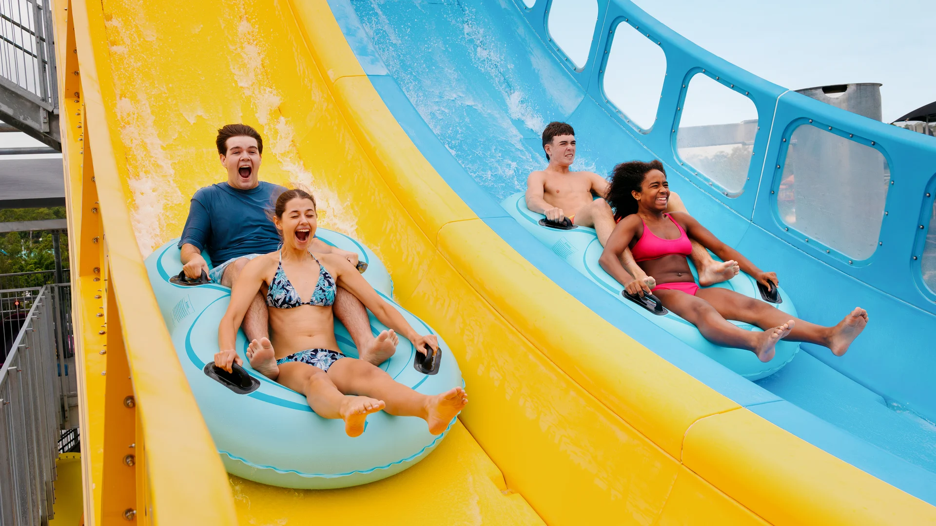 Two pairs of people in swimsuits ride inflatable rafts down bright yellow and blue water slides, smiling and laughing as they enjoy the ride on a sunny day.