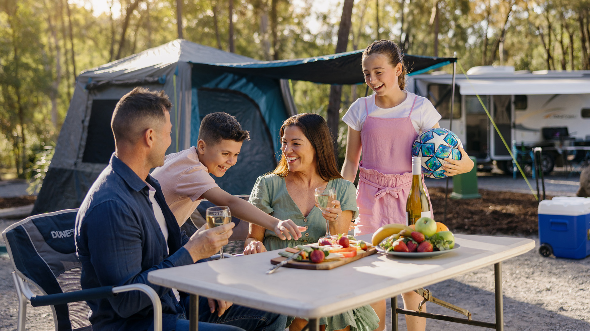 A family sits at a table outdoors near a tent, sharing food and drinks. Two adults and two children are smiling; one child holds a soccer ball. Fruit and snacks are on the table.