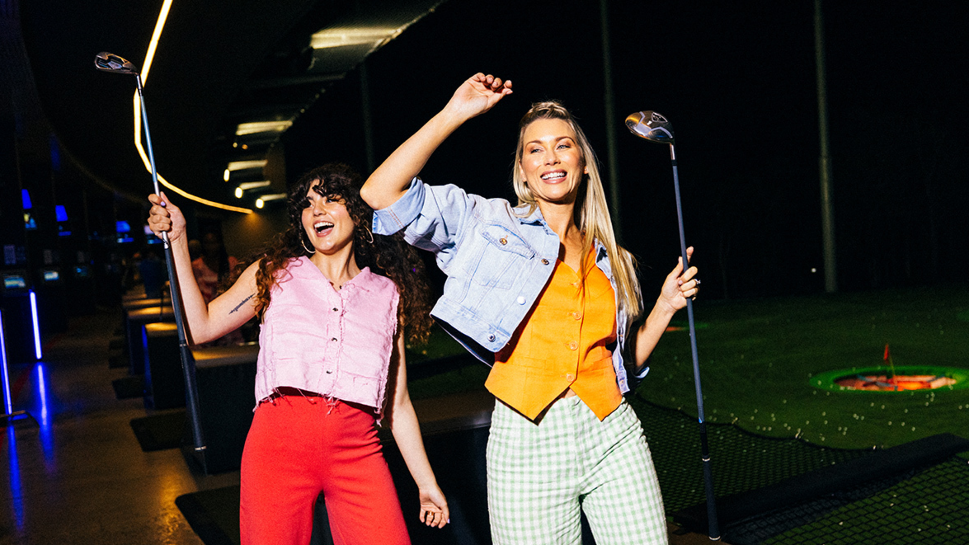 Two women are smiling and holding golf clubs in a nighttime indoor driving range, wearing colourful clothing.