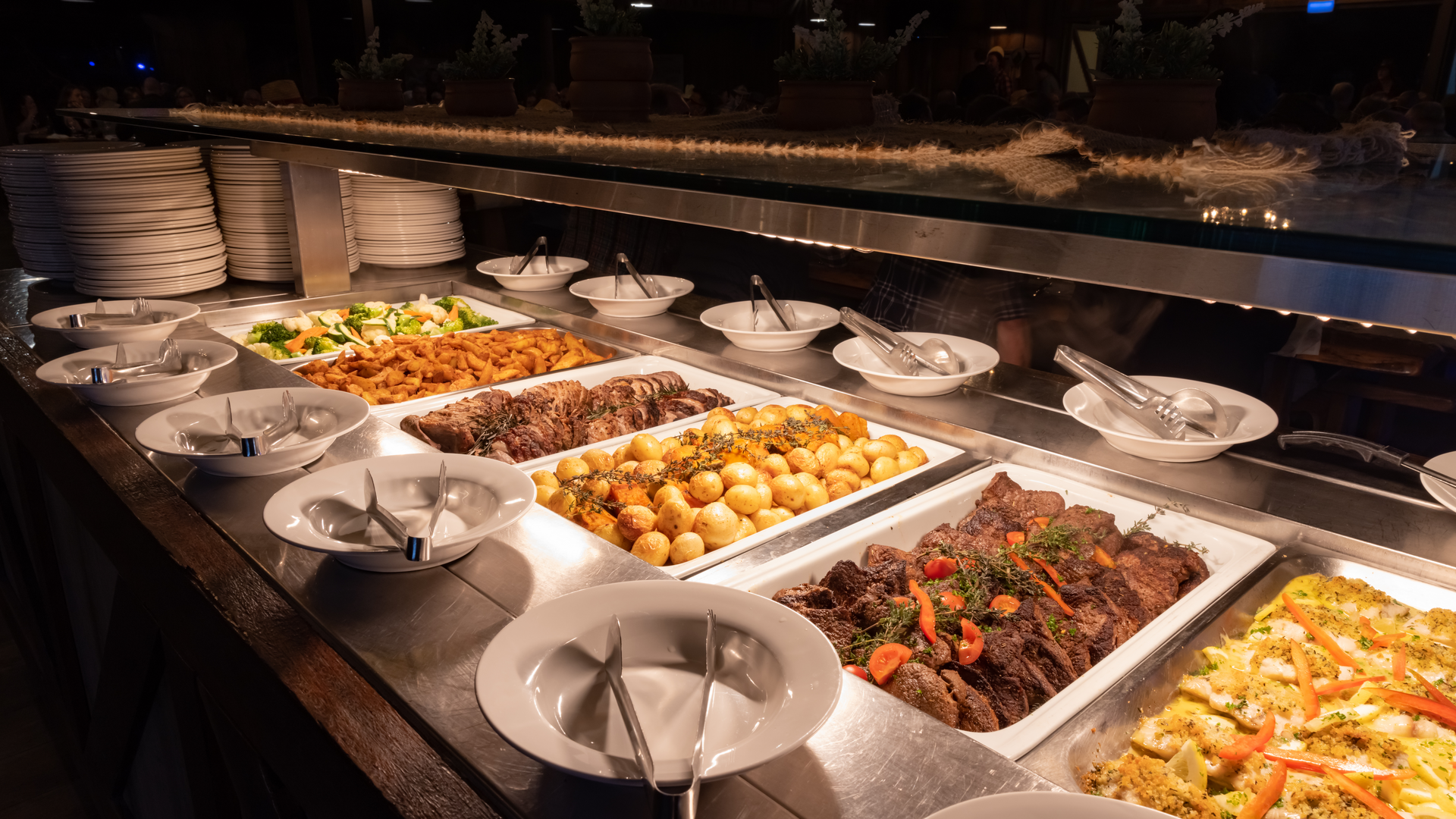 Buffet with several trays of hot food, including roasted potatoes, vegetables, and meat dishes, with plates and serving utensils arranged in front.