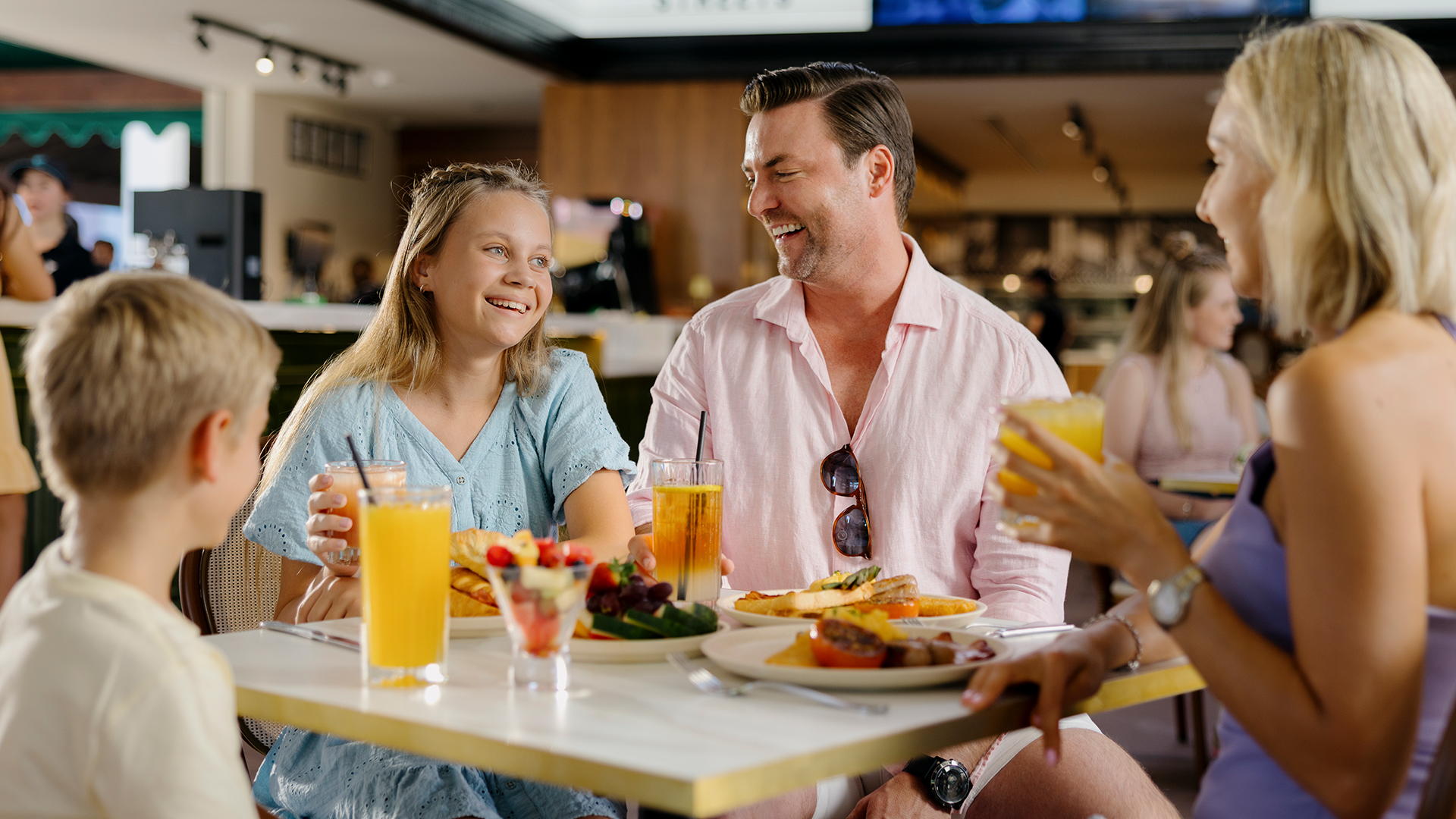A family of four sits at a restaurant table, smiling and enjoying a meal together. They have plates of food and glasses of orange juice in front of them, creating a cheerful and relaxed atmosphere.