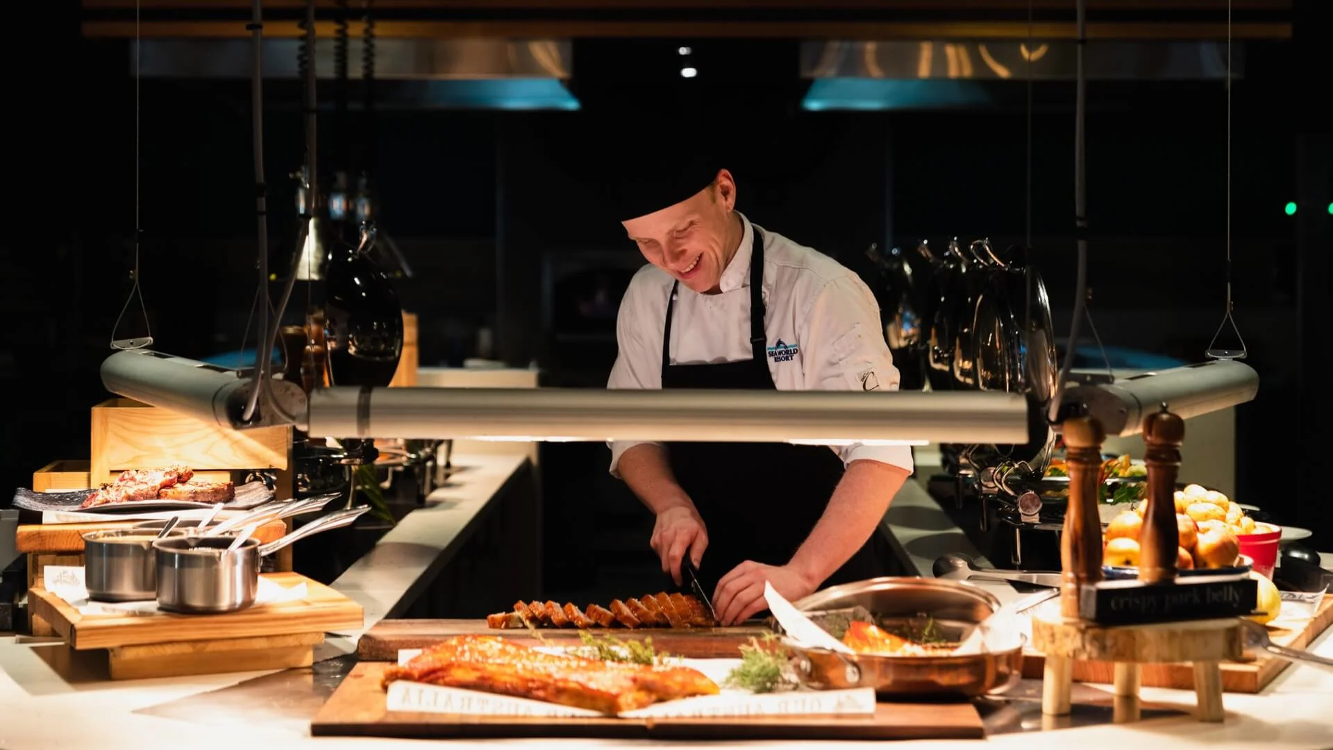 Chef at Shoreline Restaurant slices meat on a wooden board. Plates of food and small pots are arranged around him under hanging lamps.