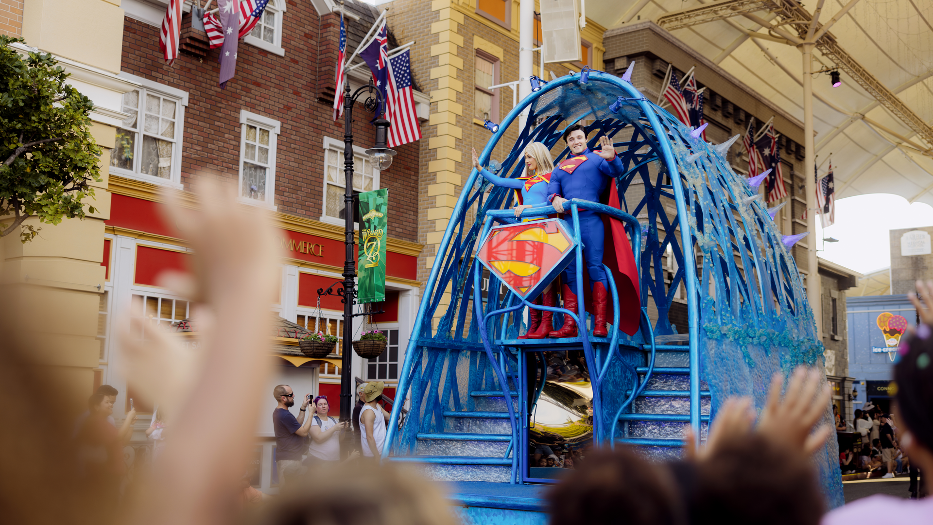 Superhero characters on a blue float wave to a crowd during a parade, with buildings and flags in the background.