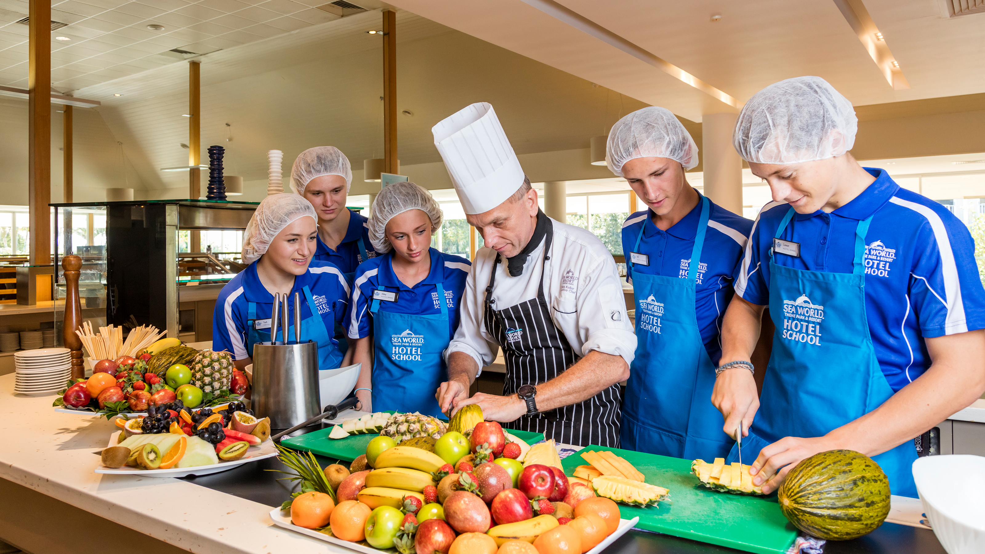 Chef instructs five students in blue uniforms on fruit preparation in a kitchen. Various fruits are displayed on the counter.