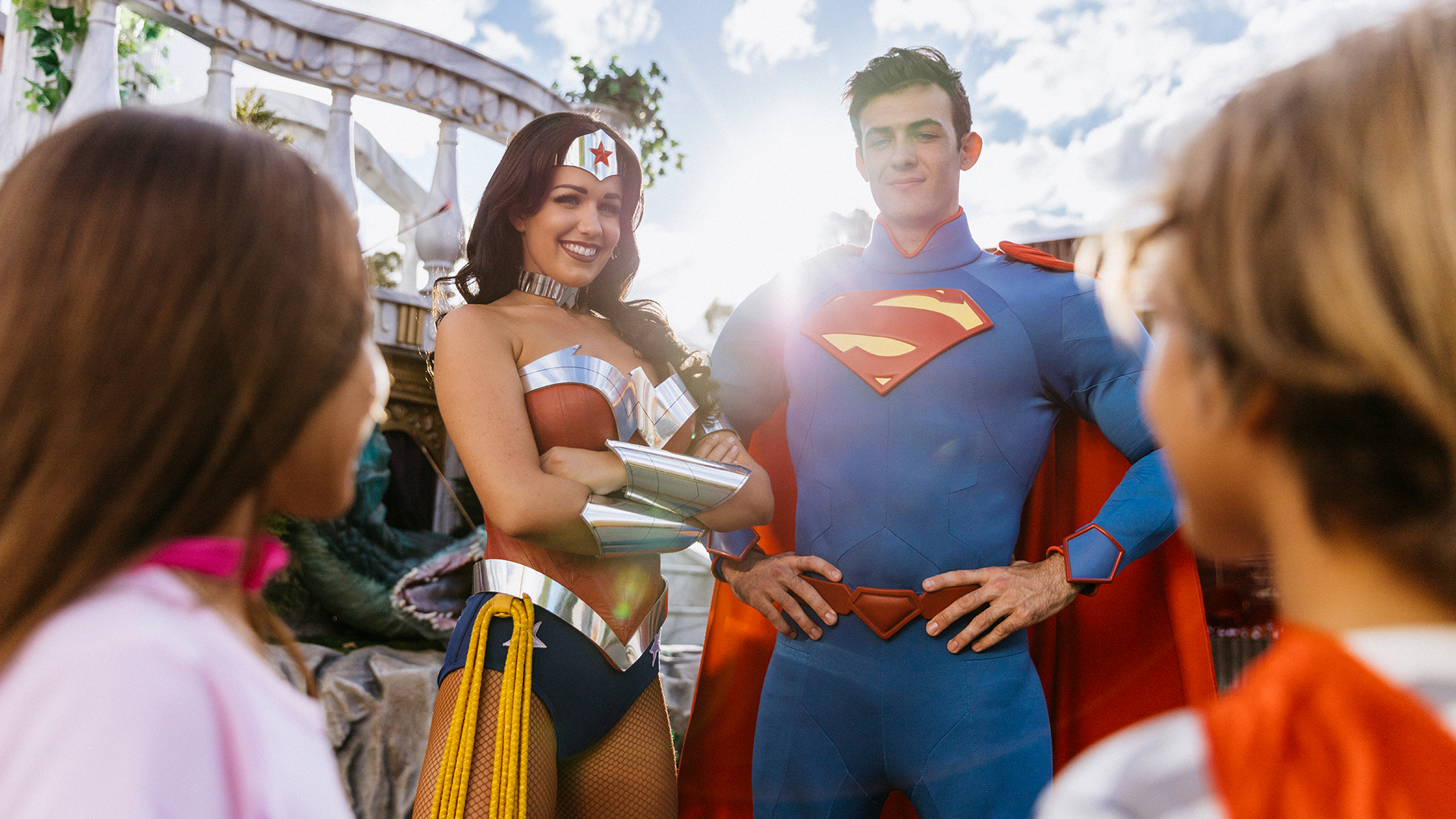 Two adults dressed as Wonder Woman and Superman stand outdoors, smiling and interacting with two children in costume, with sunlight in the background.