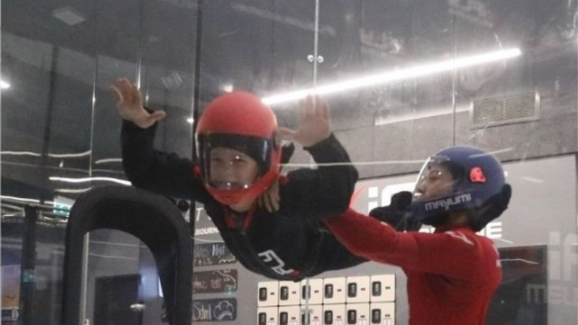 A child wearing a red helmet and flight suit is suspended in the air inside an indoor skydiving wind tunnel, assisted by an instructor in a blue helmet and red suit.
