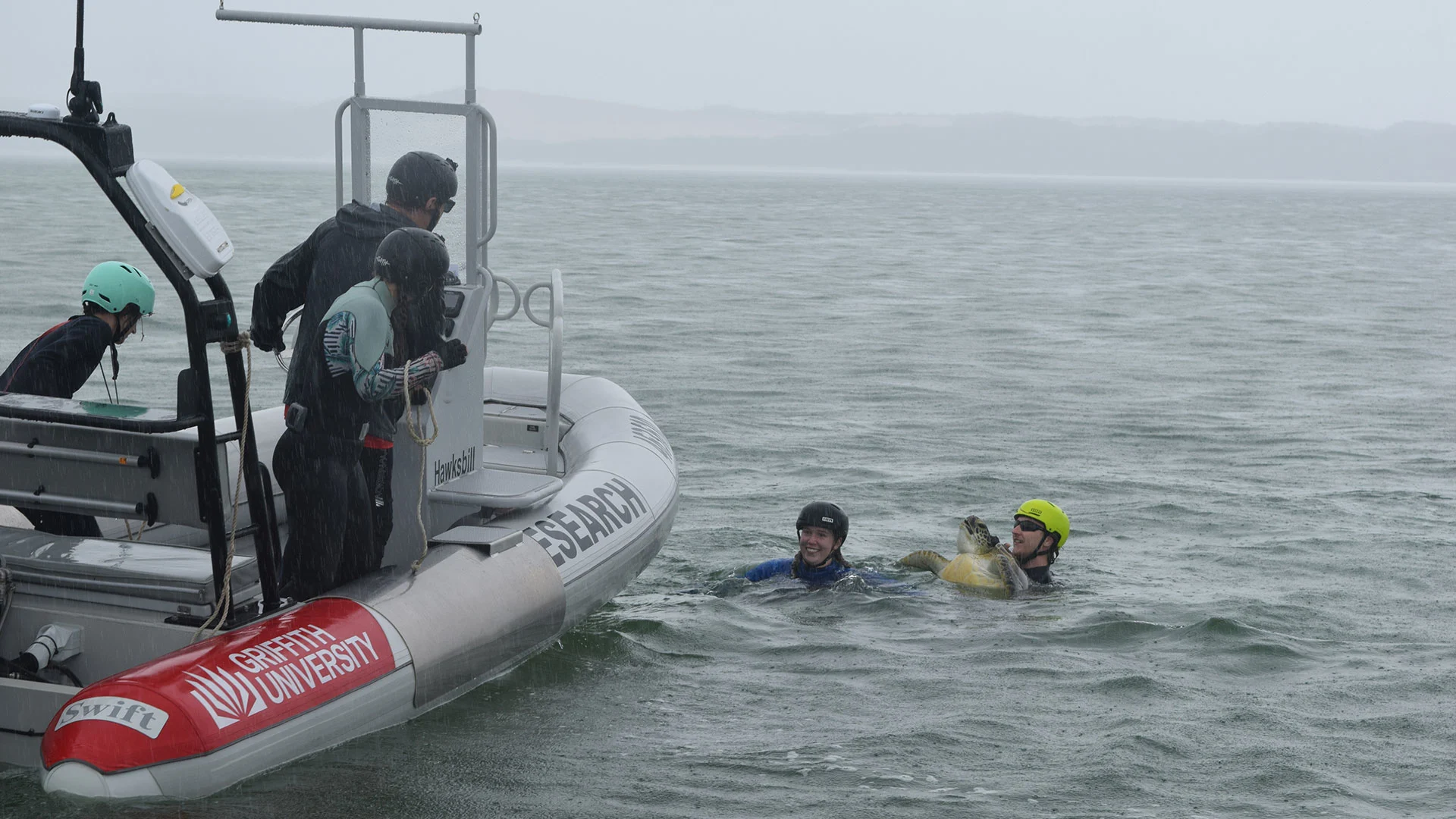 Two people in the water near a small research boat on a cloudy day, with crew members observing.