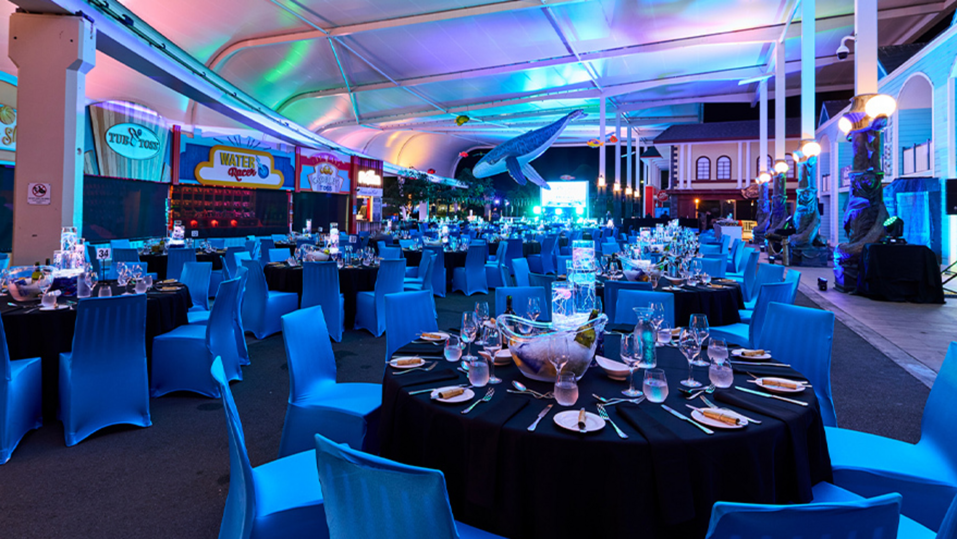 A large, colorful event space with round tables covered in black tablecloths and blue chairs, set for dinner. The ceiling is lit with blue and purple lights, and a large whale model hangs overhead.