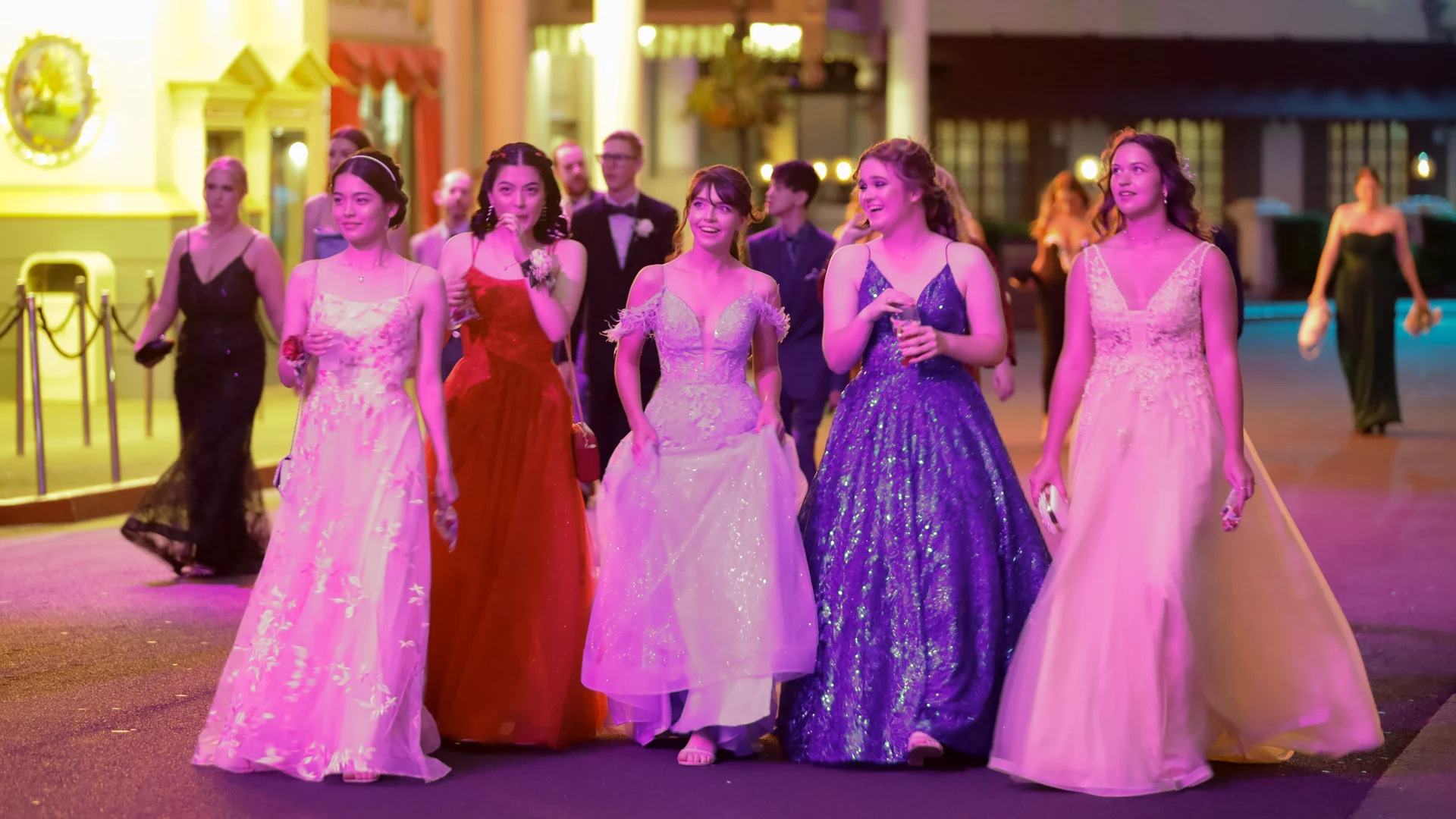 A group of young people dressed in formal gowns and suits walk together outside at night, smiling and talking, as colorful lights illuminate the scene, suggesting a special event or prom.
