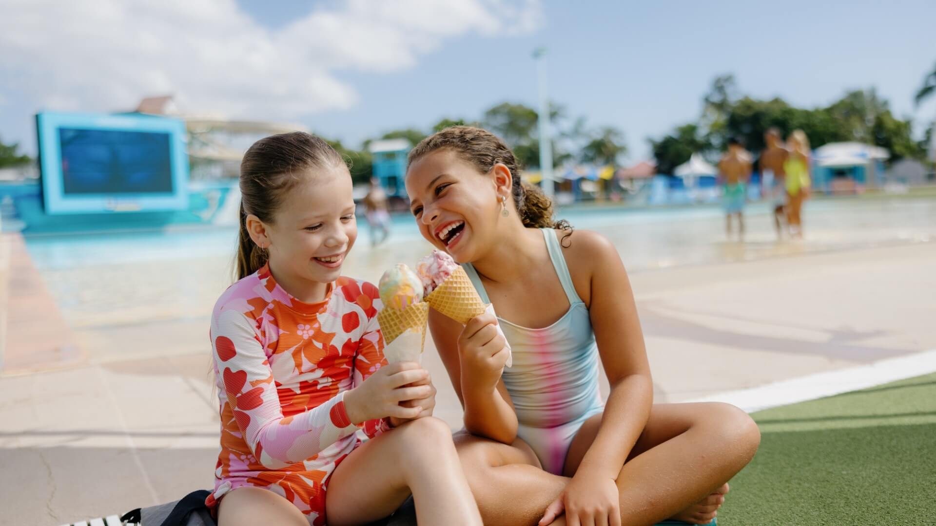 Two children sit by the Giant Wave Pool at Wet'n'Wild Water Park on the Gold Coast, wearing colourful swimsuits and happily eating ice cream cones on a sunny day.
