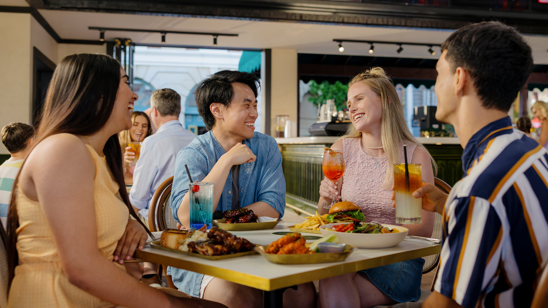 Four young adults sit at a restaurant table outdoors, laughing and enjoying drinks and a meal together. Plates of food and colorful beverages are on the table, creating a lively and cheerful atmosphere.