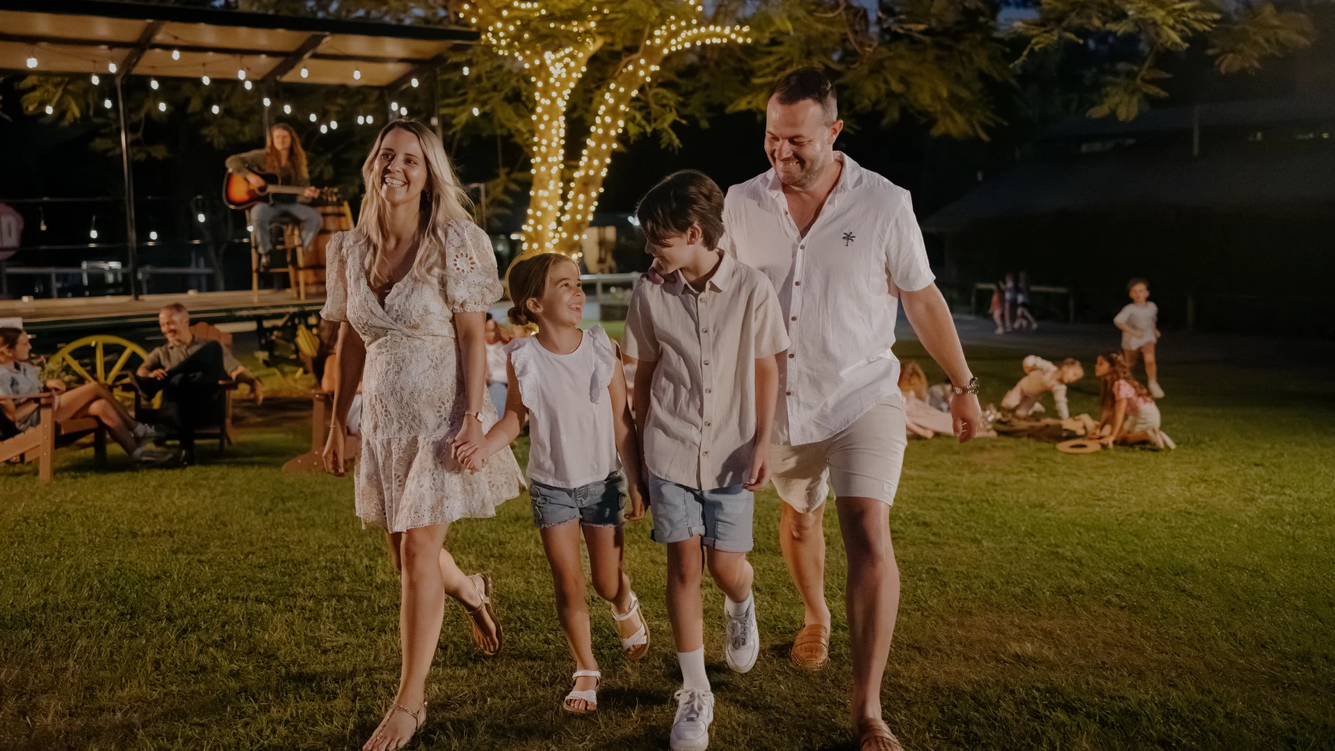 A smiling family of four, dressed in light summer clothes, walks hand in hand on grass at an outdoor event in the evening, with string lights and a musician performing on a stage in the background.