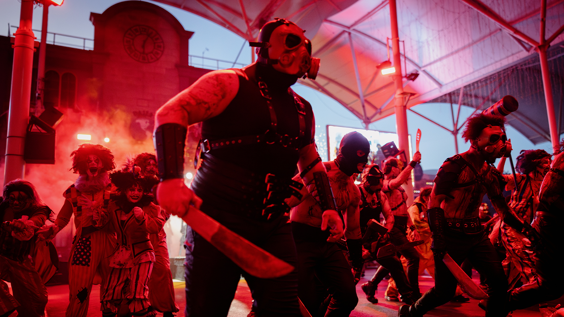 A group of people in dark, creepy costumes and masks, some holding machetes, participate in a dramatic, red-lit scene under a metal-roofed structure with smoke in the background.