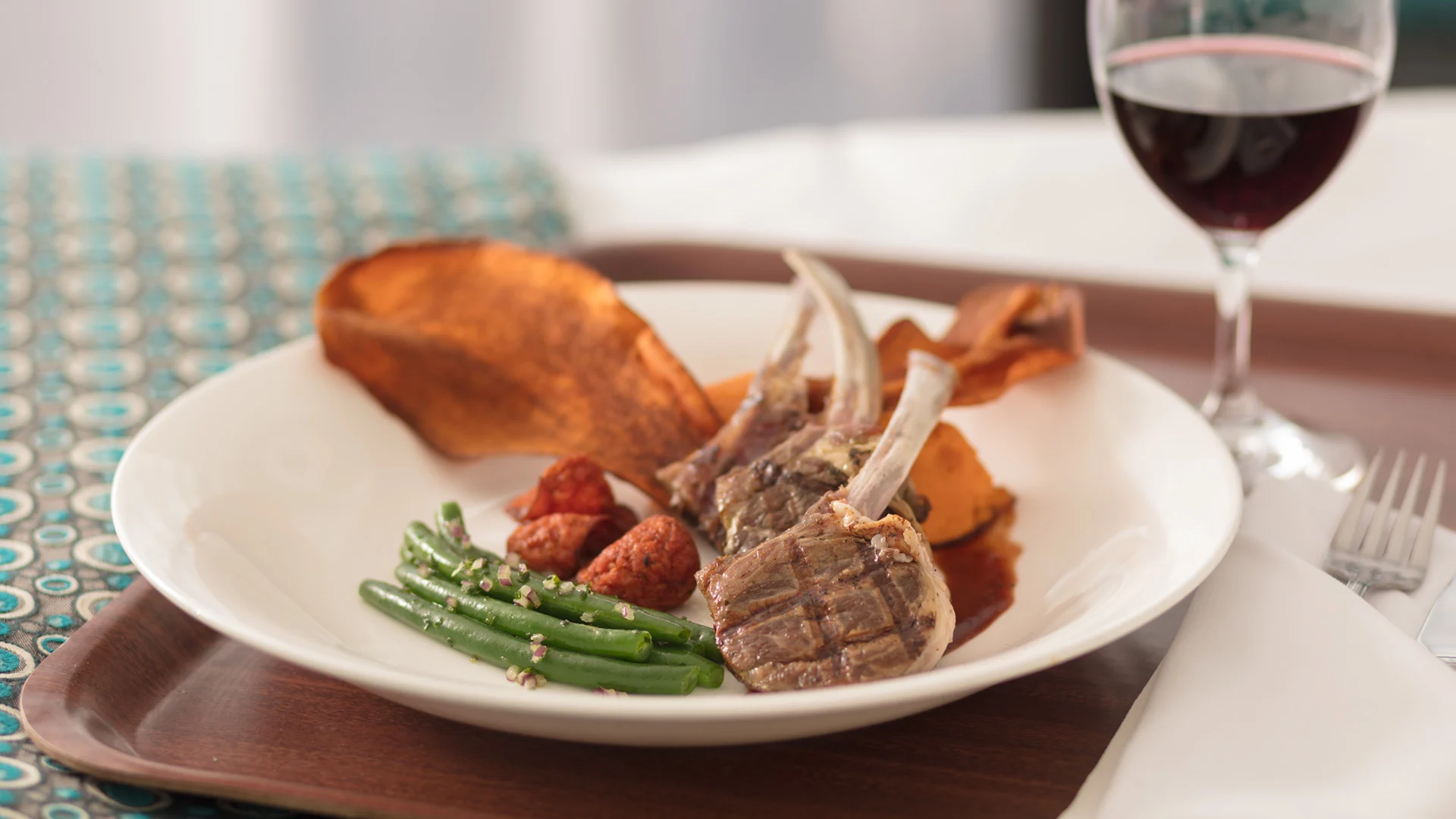 Plate of grilled lamb chops with green beans, roasted potatoes, and a sweet potato slice, accompanied by a glass of red wine.