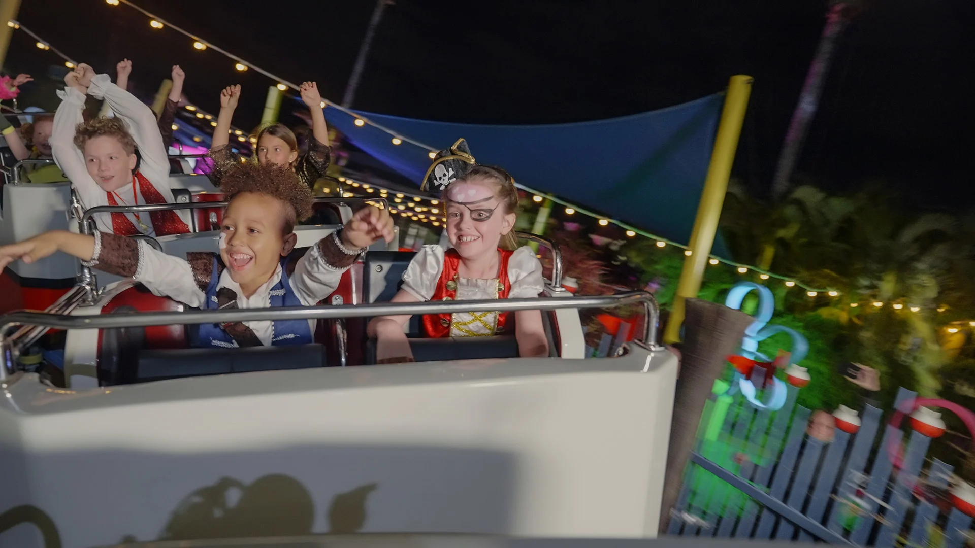 Children dressed as pirates ride a roller coaster at night, smiling and raising their arms in excitement. String lights and greenery are visible in the festive background.