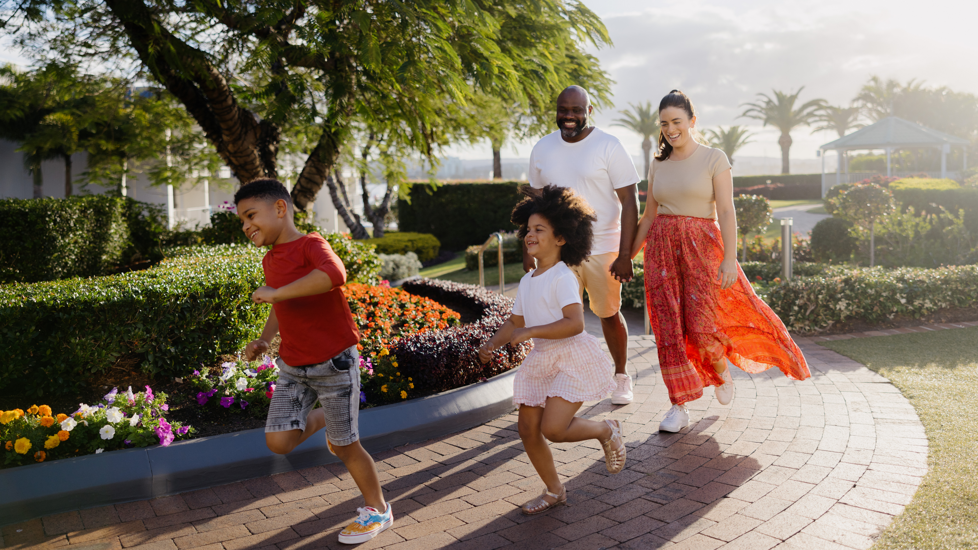 A family of four walks along a brick path at Sea World Resort. Two children run ahead while the parents follow, smiling. The background features bushes, flowers, and trees.