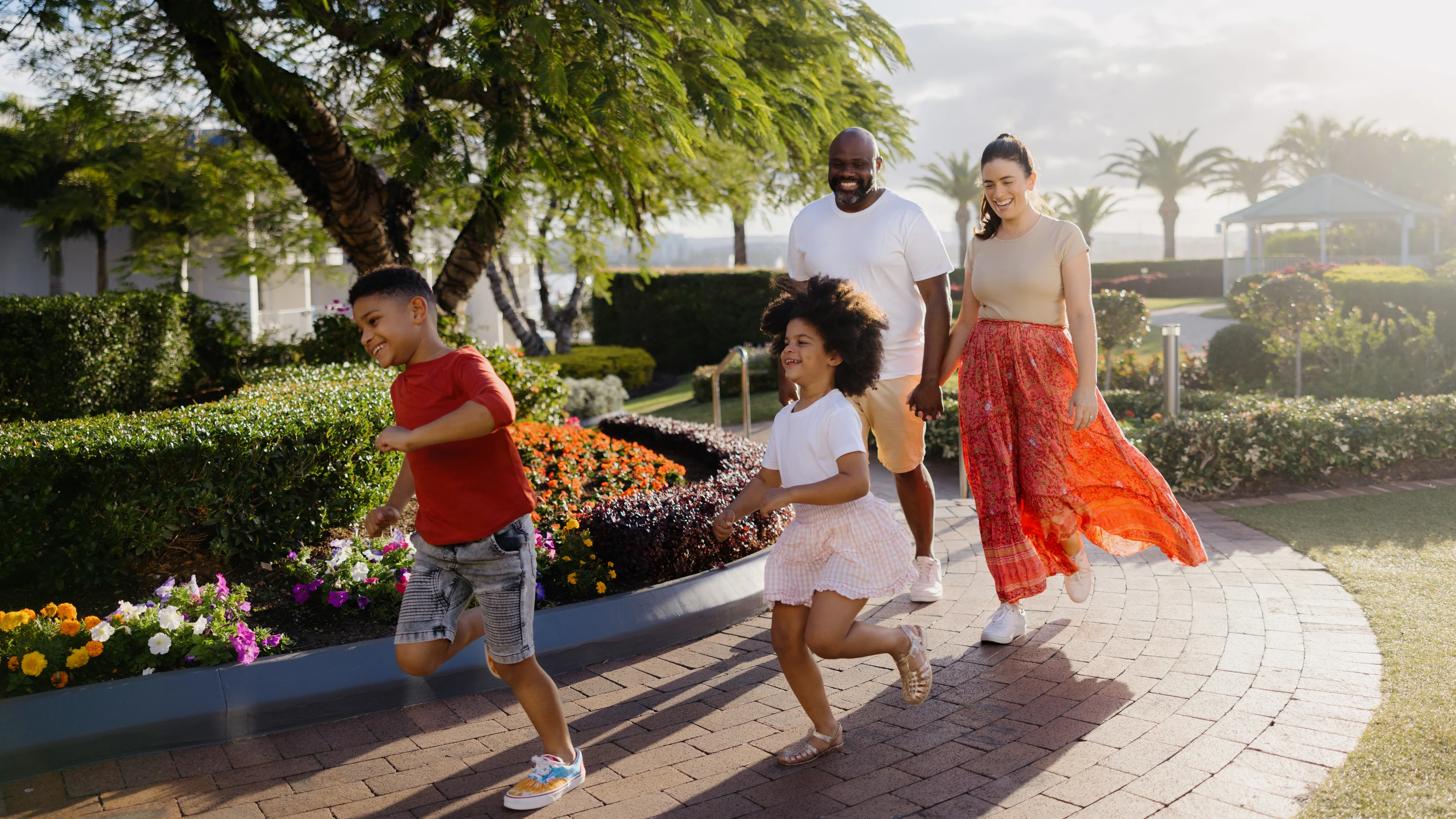 A family of four walks along a brick path at Sea World Resort. Two children run ahead while the parents follow, smiling. The background features bushes, flowers, and trees.