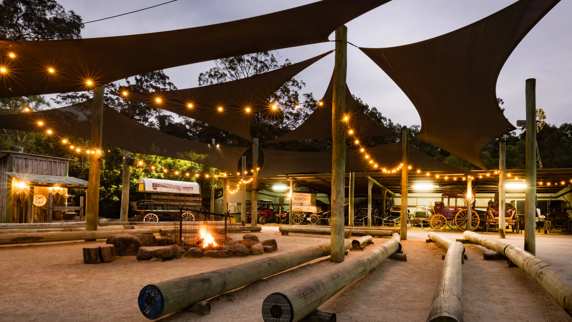 Outdoor area with string lights, a central fire pit, wooden logs for seating, sunshades overhead, and rustic buildings in the background.