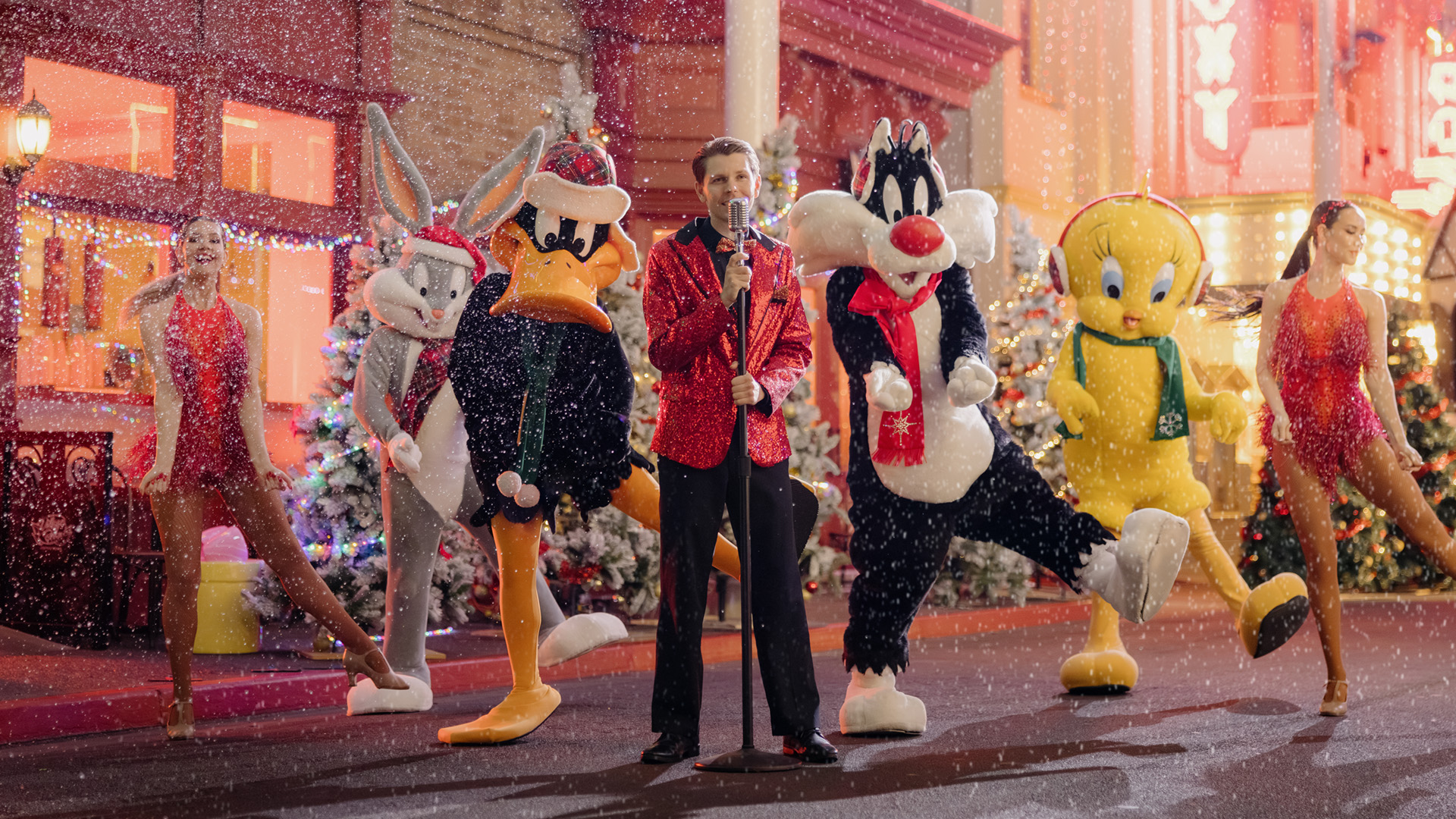 A performer in a red jacket sings on a snowy street, surrounded by dancers and people in Bugs Bunny, Daffy Duck, Sylvester, and Tweety Bird costumes, with Christmas trees and festive lights in the background.