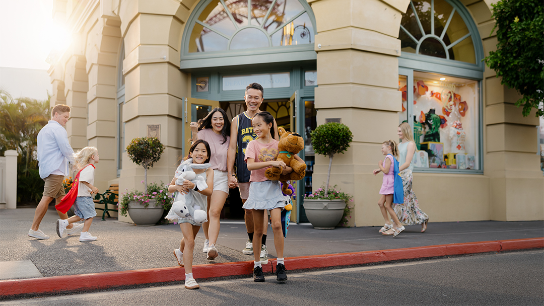 A smiling family with two children carrying stuffed animals walks out of a toy store, while other people stroll nearby on a sunny day.