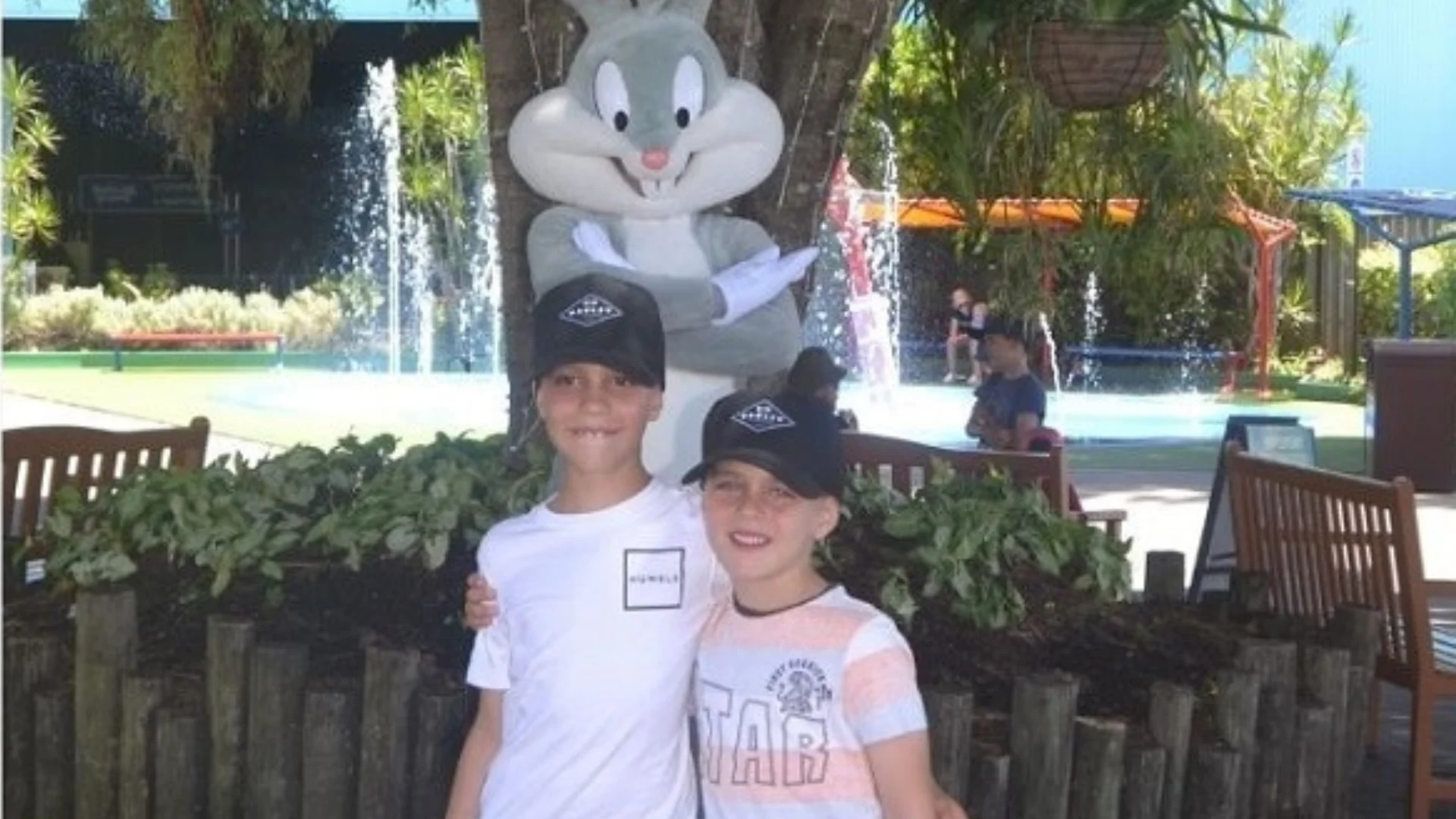 Two children wearing black hats pose and smile in front of a large Bugs Bunny statue, with a tree, plants, a fountain, and some outdoor seating in the background.