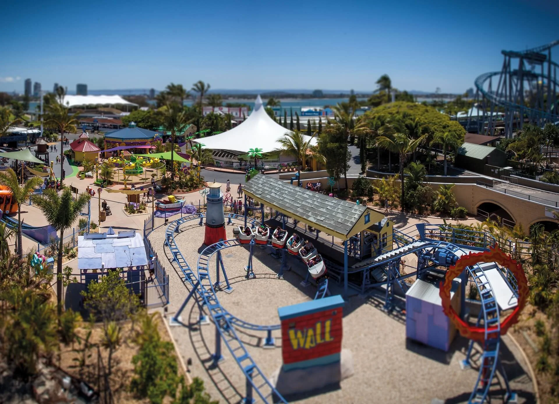 An amusement park with various rides, roller coasters, and colorful attractions under a clear blue sky, surrounded by trees and distant water.