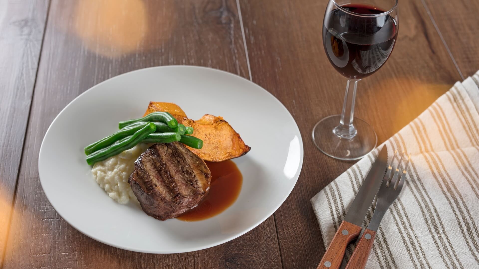 Plate of grilled steak with green beans, roasted pumpkin, and risotto, served with a glass of red wine on a wooden table