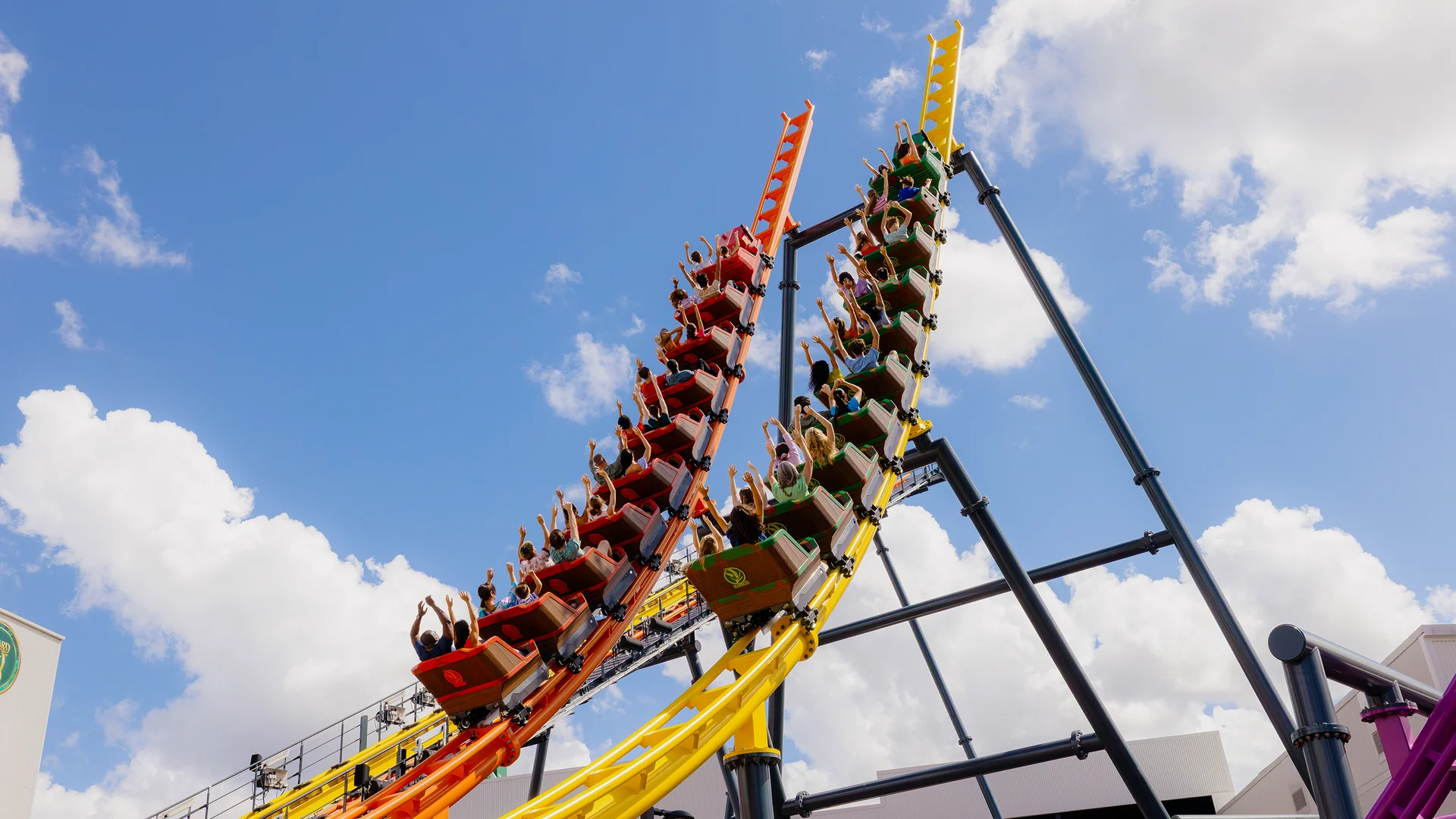 Two roller coaster cars, one orange and one yellow, filled with people holding their hands up, ascend steep parallel tracks against a bright blue sky with scattered clouds.