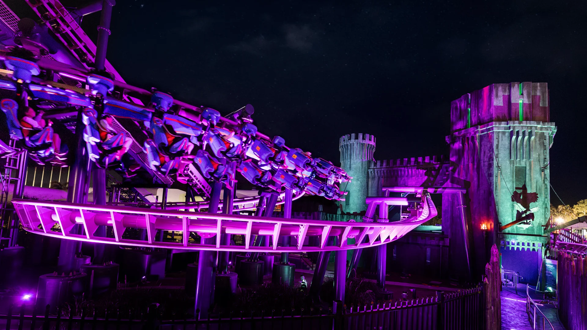 A roller coaster train speeds through a sharp turn at night, illuminated by vibrant purple and green lights, with castle-like structures in the background.